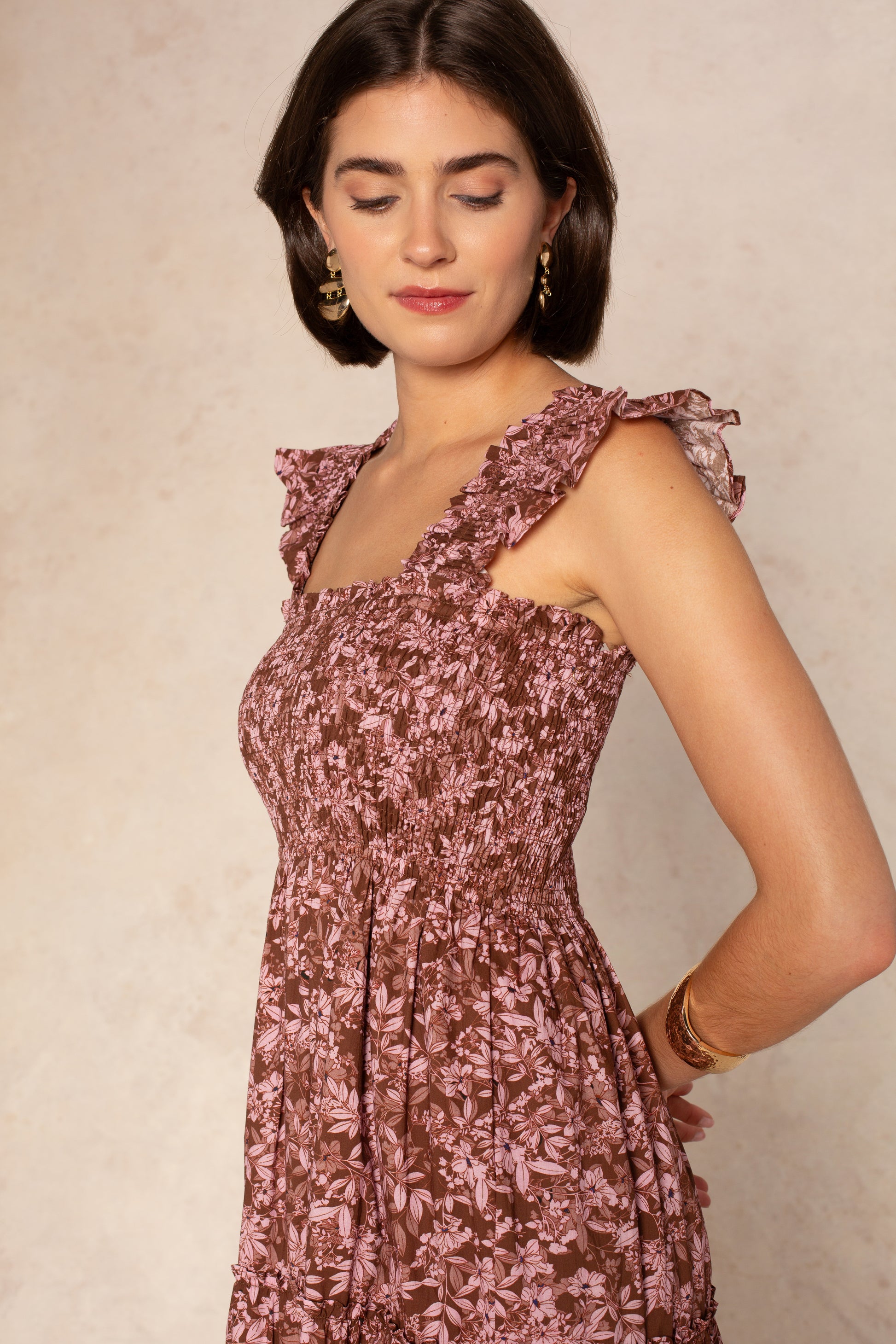 A woman with short dark hair and gold earrings wears the Georgie Calypso Smocked Midi Dress, featuring ruffled straps and a floral print, as she stands against a light background looking down with a gentle expression.