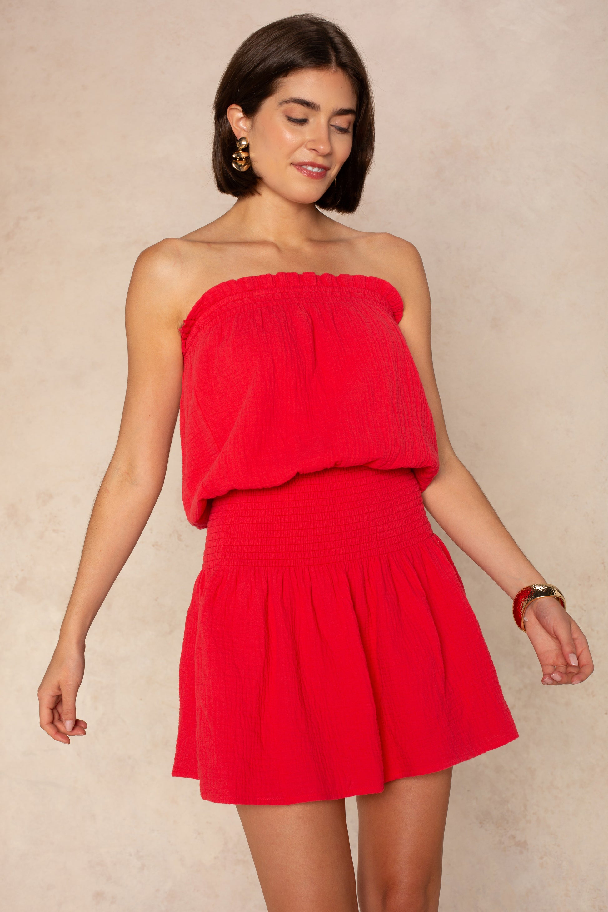 A woman with short brown hair wears the Isla Mila Strapless Dress Coverup—a bright red, textured mini perfect for the beach or as a swim coverup. She pairs it with gold hoop earrings and a bracelet, smiling gently against a light beige background.