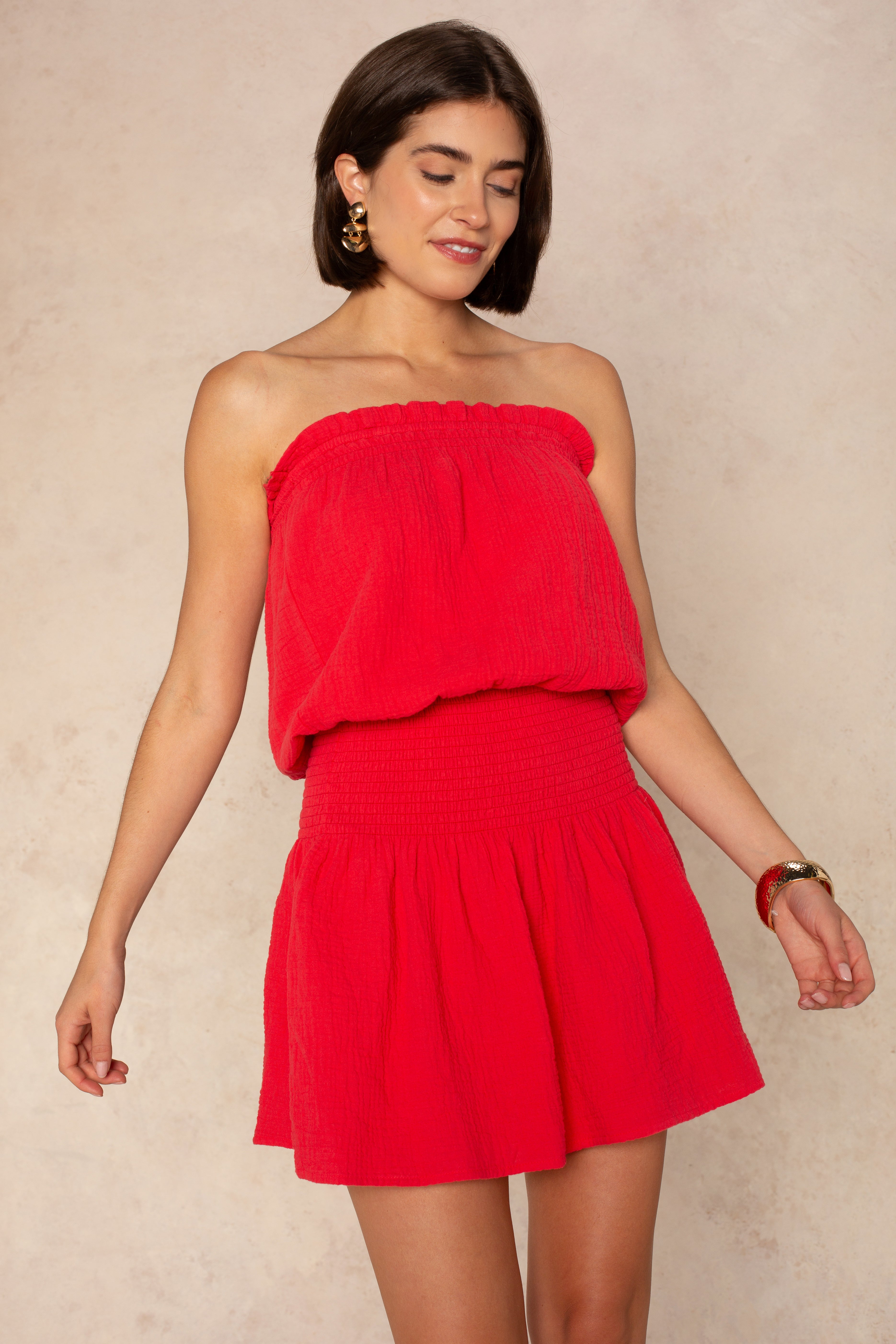 A woman with short brown hair wears the Isla Mila Strapless Dress Coverup—a bright red, textured mini perfect for the beach or as a swim coverup. She pairs it with gold hoop earrings and a bracelet, smiling gently against a light beige background.
