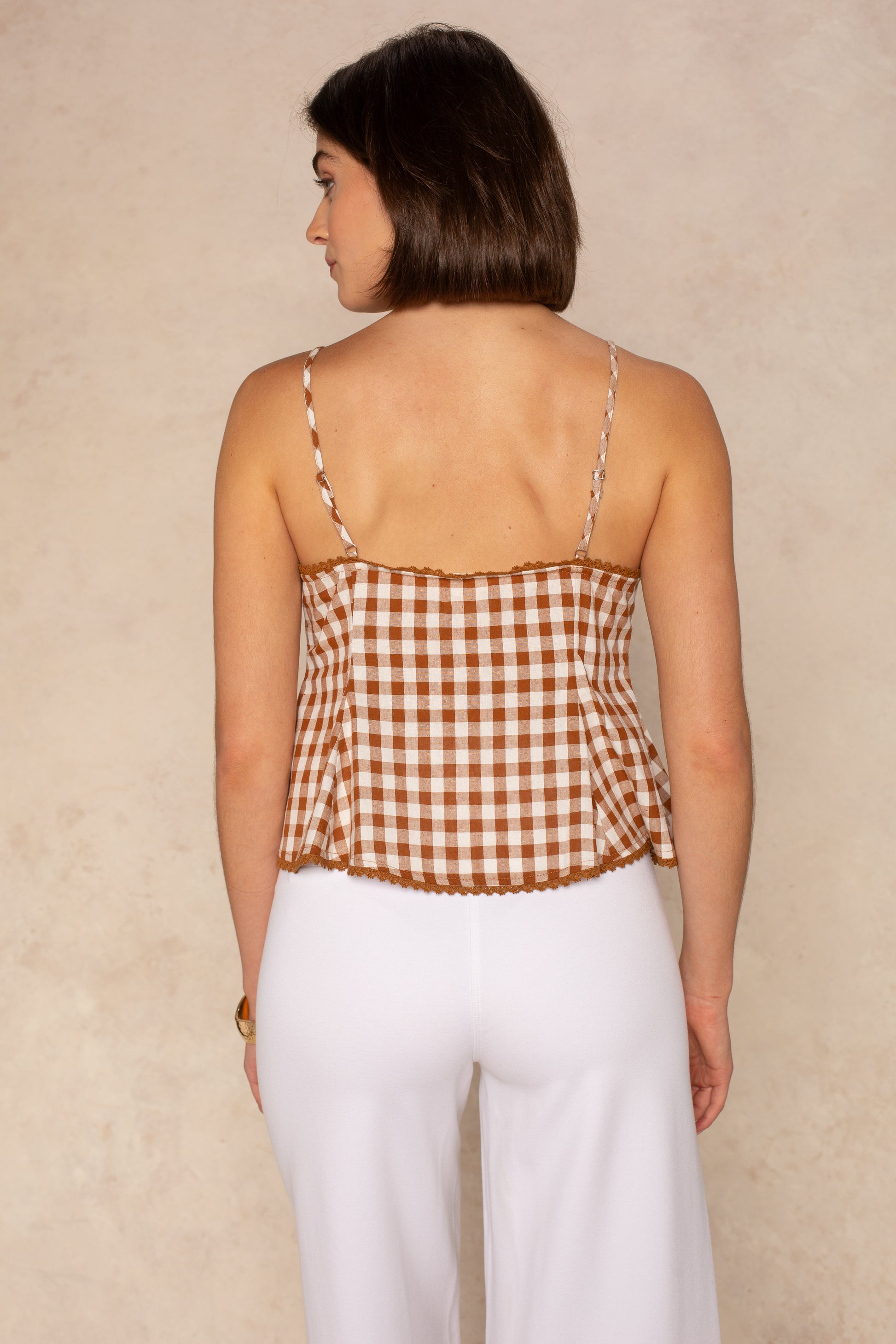 A woman with shoulder-length brown hair, seen from the back, wears the Isla Darla Gingham Tie Front Top and high-waisted white pants, standing against a light neutral background.