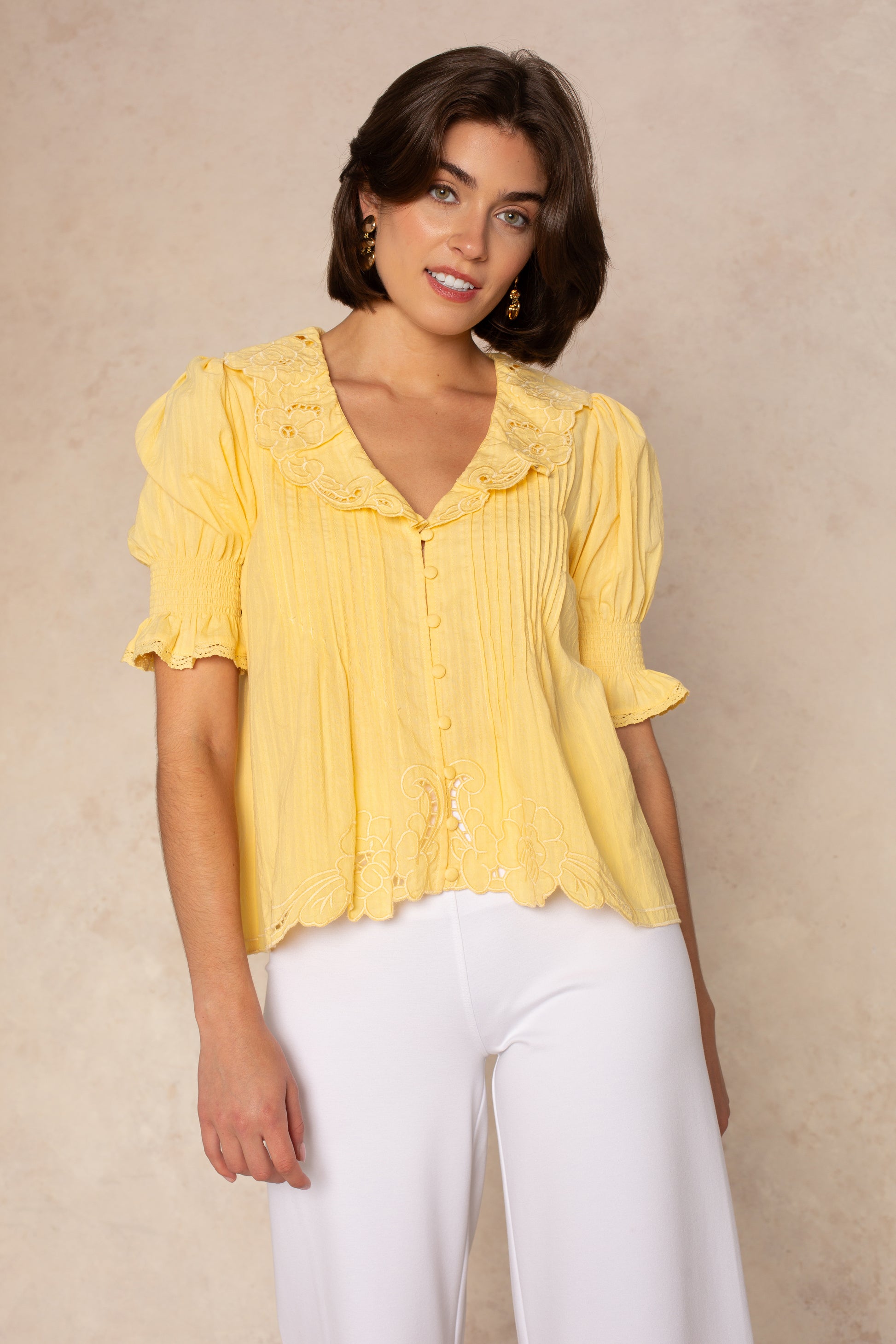 A woman with short brown hair wears the Isla Lea Embroidered Blouse—a vintage-inspired yellow top with a ruffled collar—paired with white pants, standing and smiling slightly against a beige background.