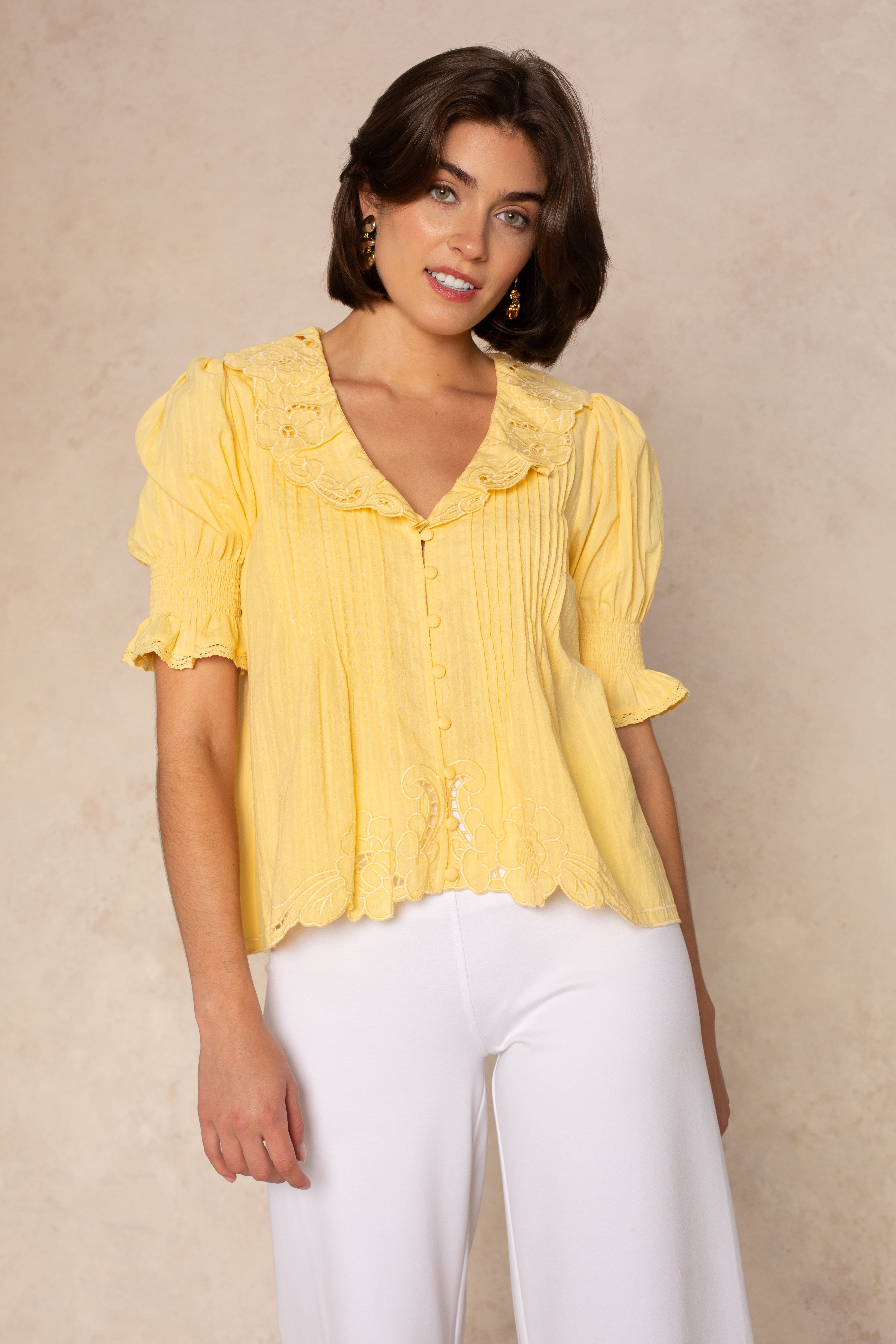 A woman with short brown hair wears the Isla Lea Embroidered Blouse—a vintage-inspired yellow top with a ruffled collar—paired with white pants, standing and smiling slightly against a beige background.