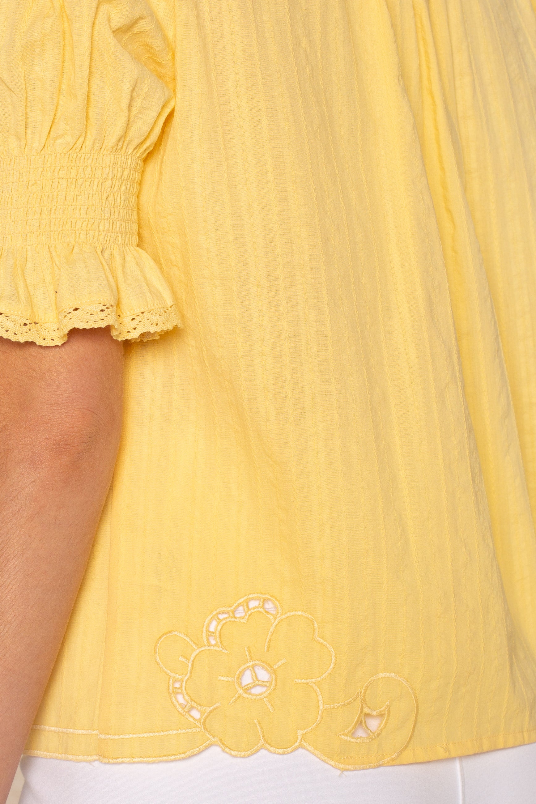 A close-up of the Lea Embroidered Blouse by Isla, a vintage-inspired yellow top with smocked puff sleeves and floral embroidery near the hem, styled over white pants.