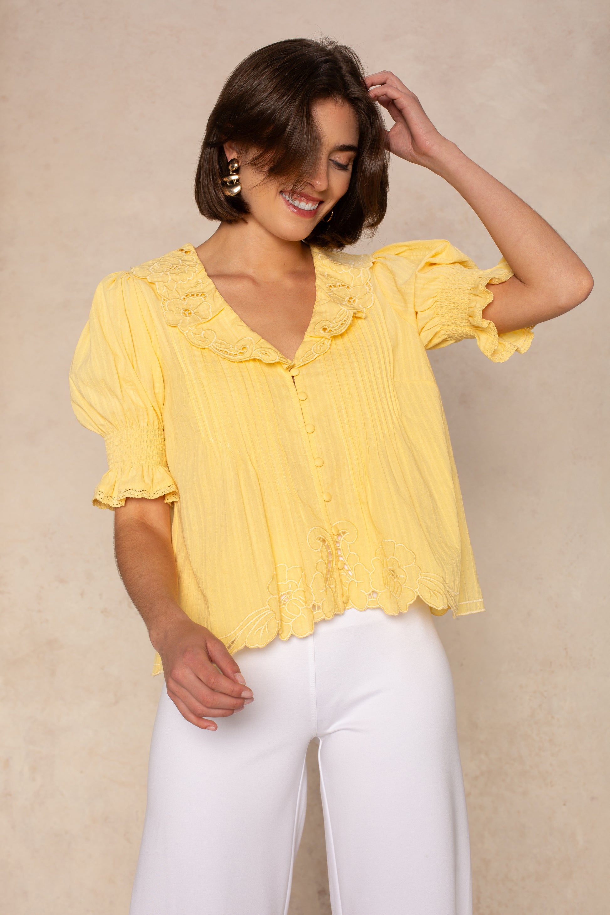 A woman with short brown hair smiles while wearing the Isla Lea Embroidered Blouse in yellow and white pants, standing against a beige background.