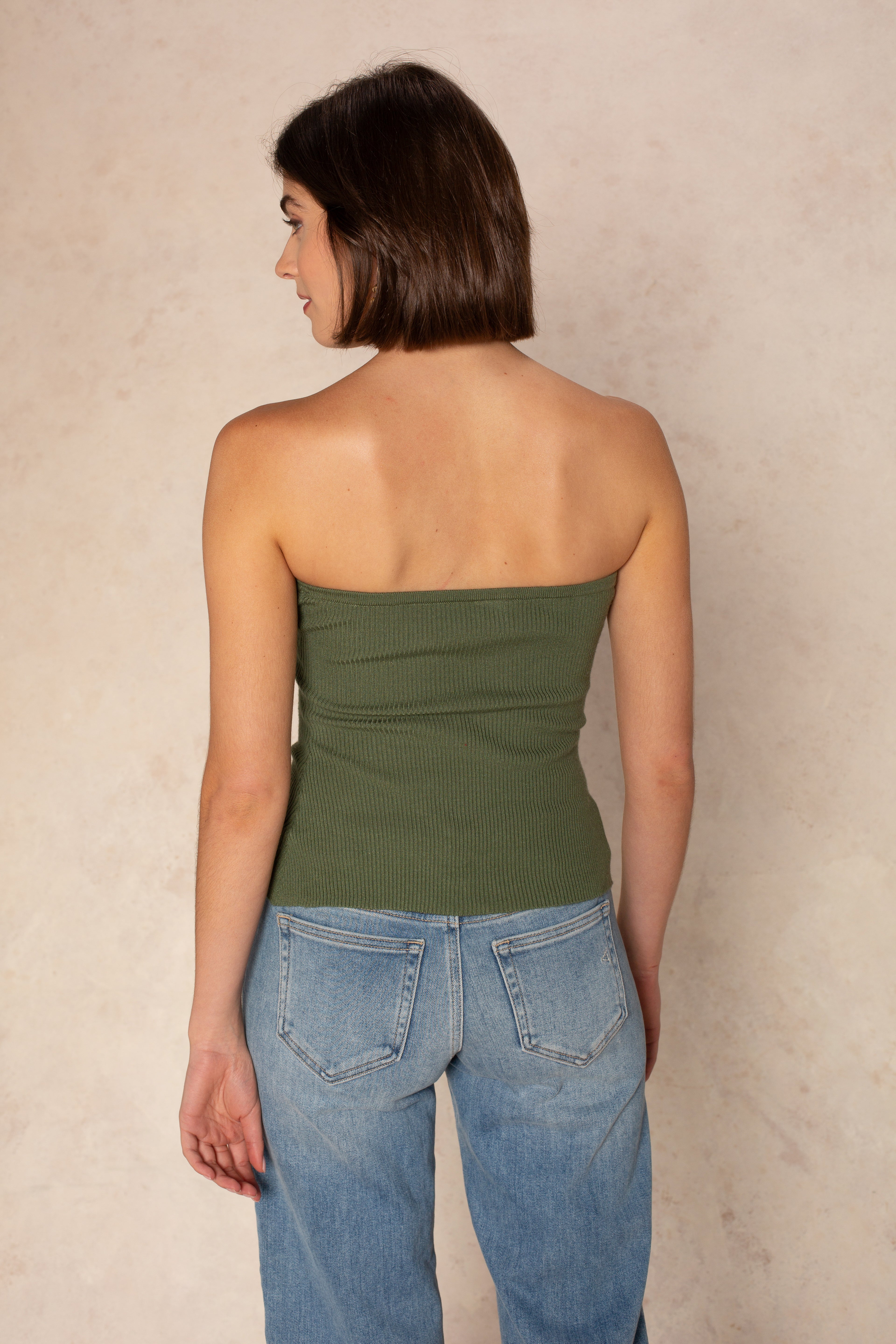A woman with short brown hair stands with her back to the camera wearing the Isla Stacy Strapless Button Front Tank in green and light blue jeans, both offering a flattering fit against a neutral, textured background.