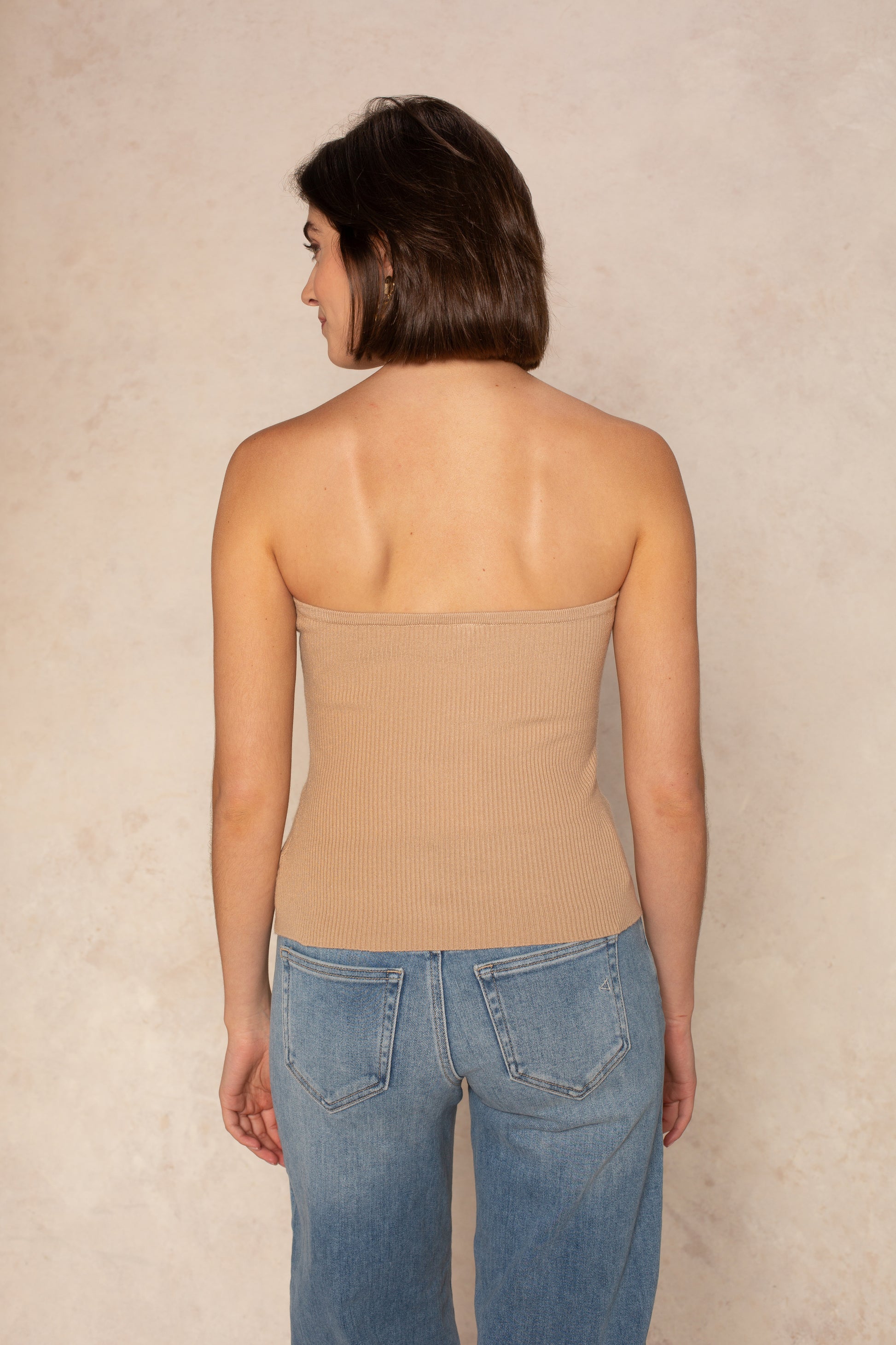 A woman with short brown hair stands with her back to the camera, wearing the Isla Stacy Strapless Button Front Tank in beige and blue jeans against a light beige textured background.