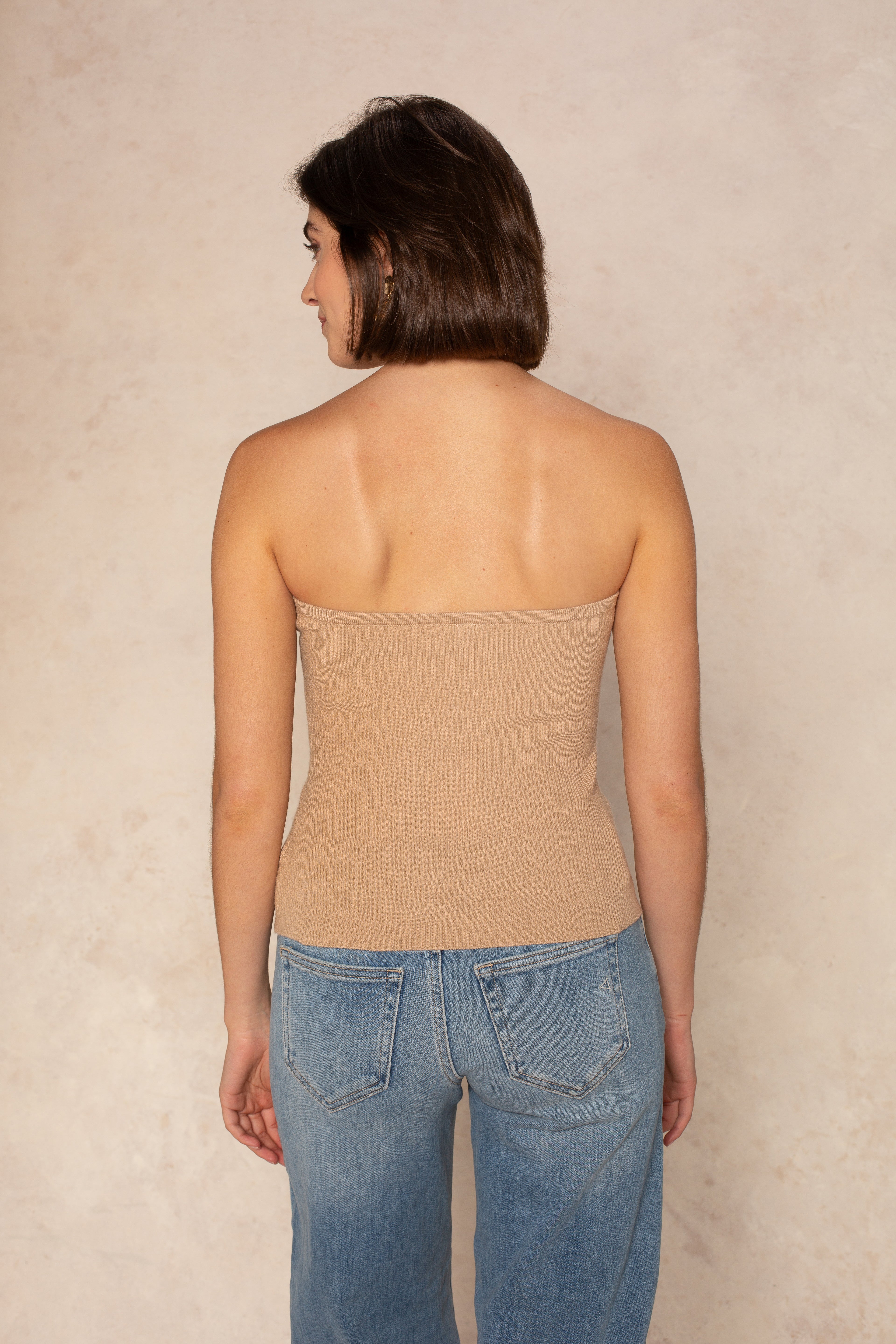 A woman with short brown hair stands with her back to the camera, wearing the Isla Stacy Strapless Button Front Tank in beige and blue jeans against a light beige textured background.