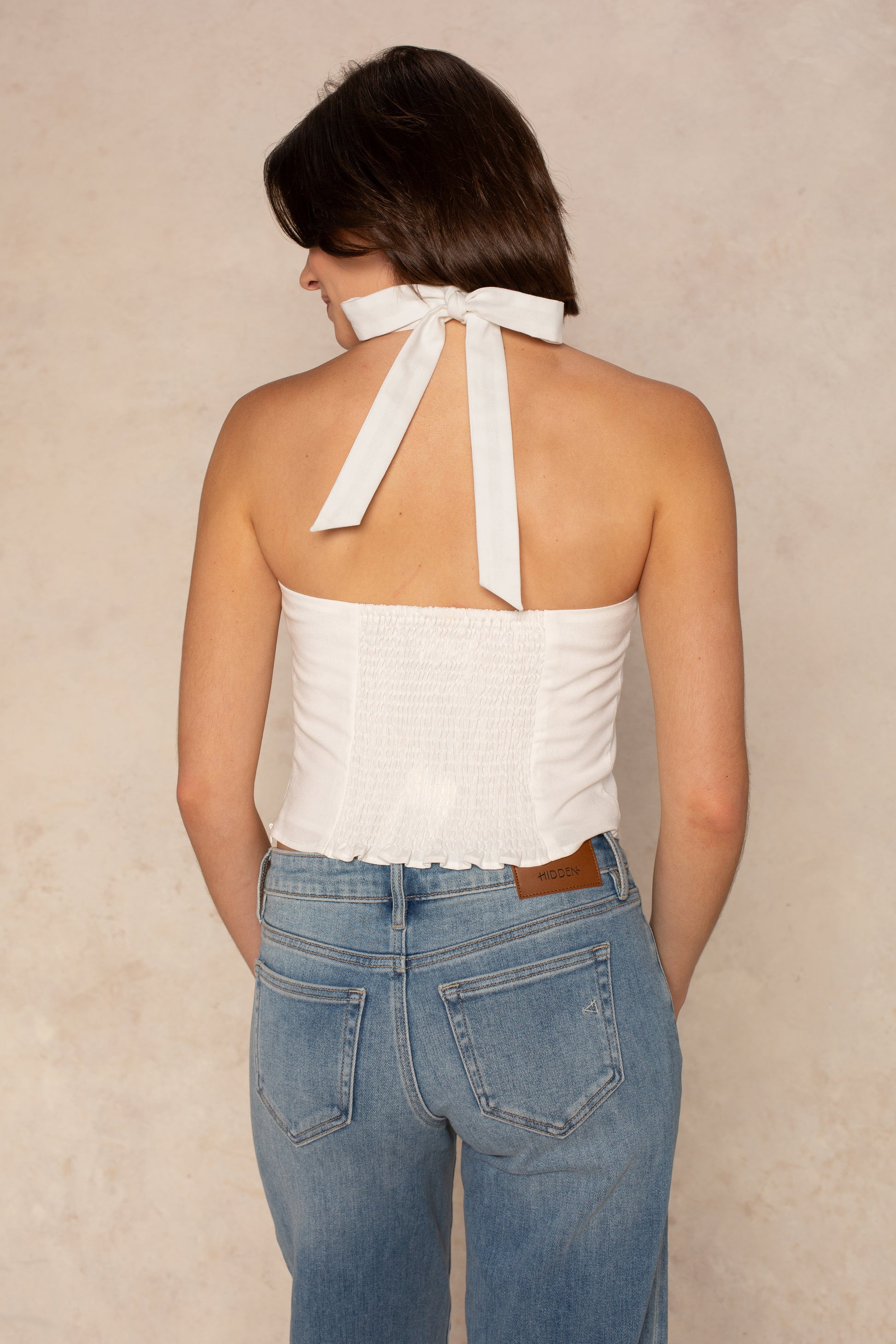 A woman stands facing away in the Georgie Harper Halter Linen Top, a white cropped tie-neck style, paired with light blue high-waisted jeans featuring a brown waistband label. The neutral background highlights the chic contrast.