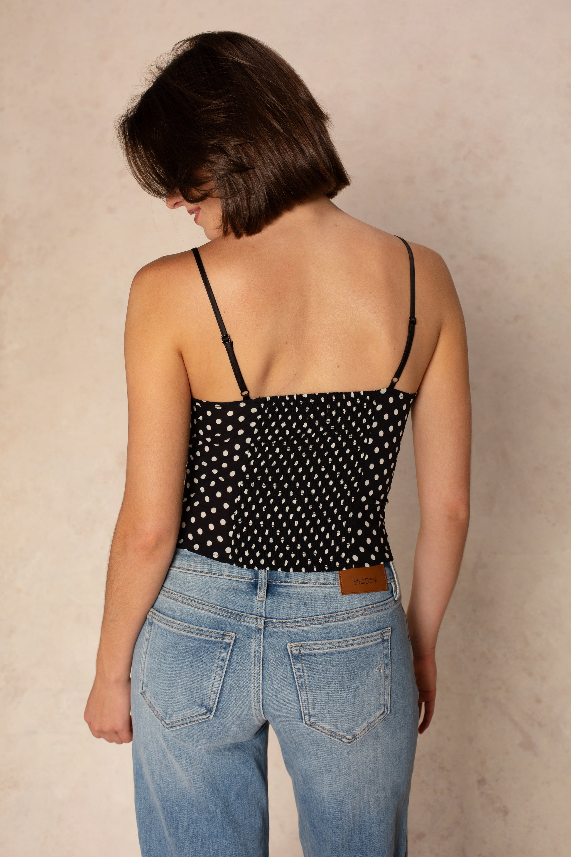 A woman with short brown hair faces away, wearing the Georgie Allison Top—a black polka dot top with a sweetheart neckline—and light blue high-waisted jeans, standing against a neutral, textured background.