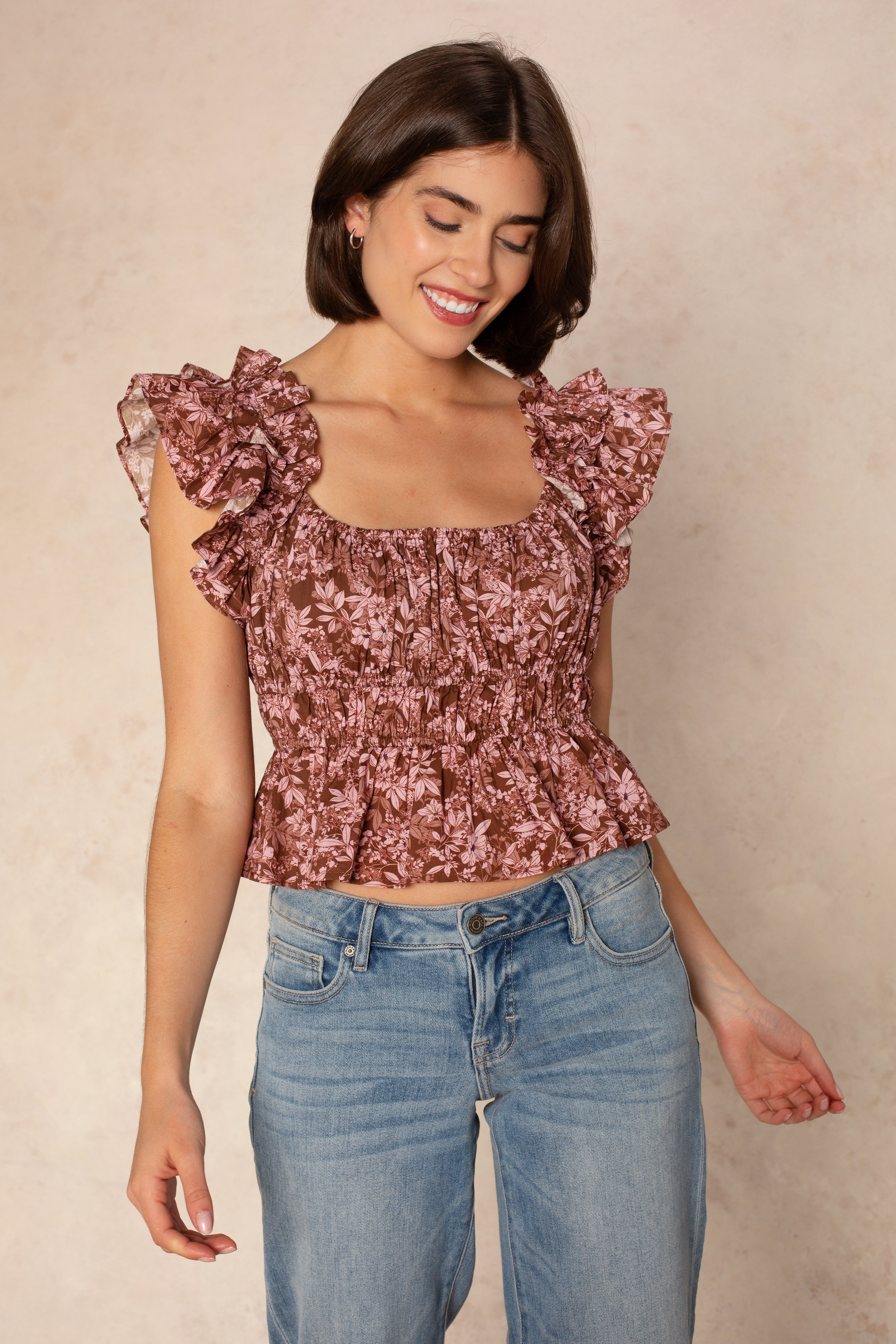 A woman with short brown hair wears the Georgie Avery Top, featuring a floral print and ruffle sleeves, paired with blue jeans. She smiles and looks down with one hand slightly lifted, standing against a light beige background.