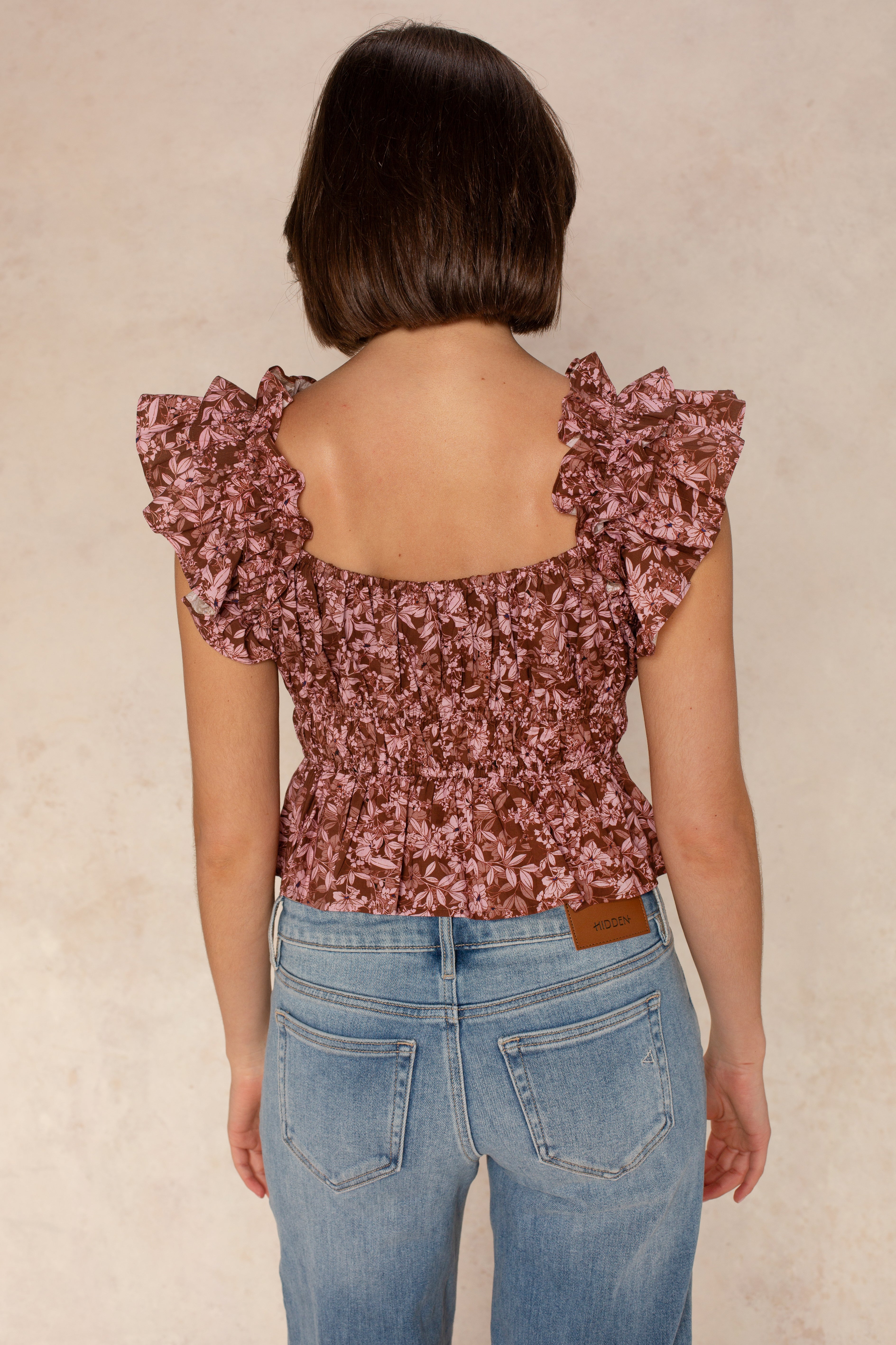 A woman with short brown hair, seen from behind, wears the Georgie Avery Top—a mauve floral print top with ruffle sleeves—and light blue high-waisted jeans with a brown leather patch on the waistband.