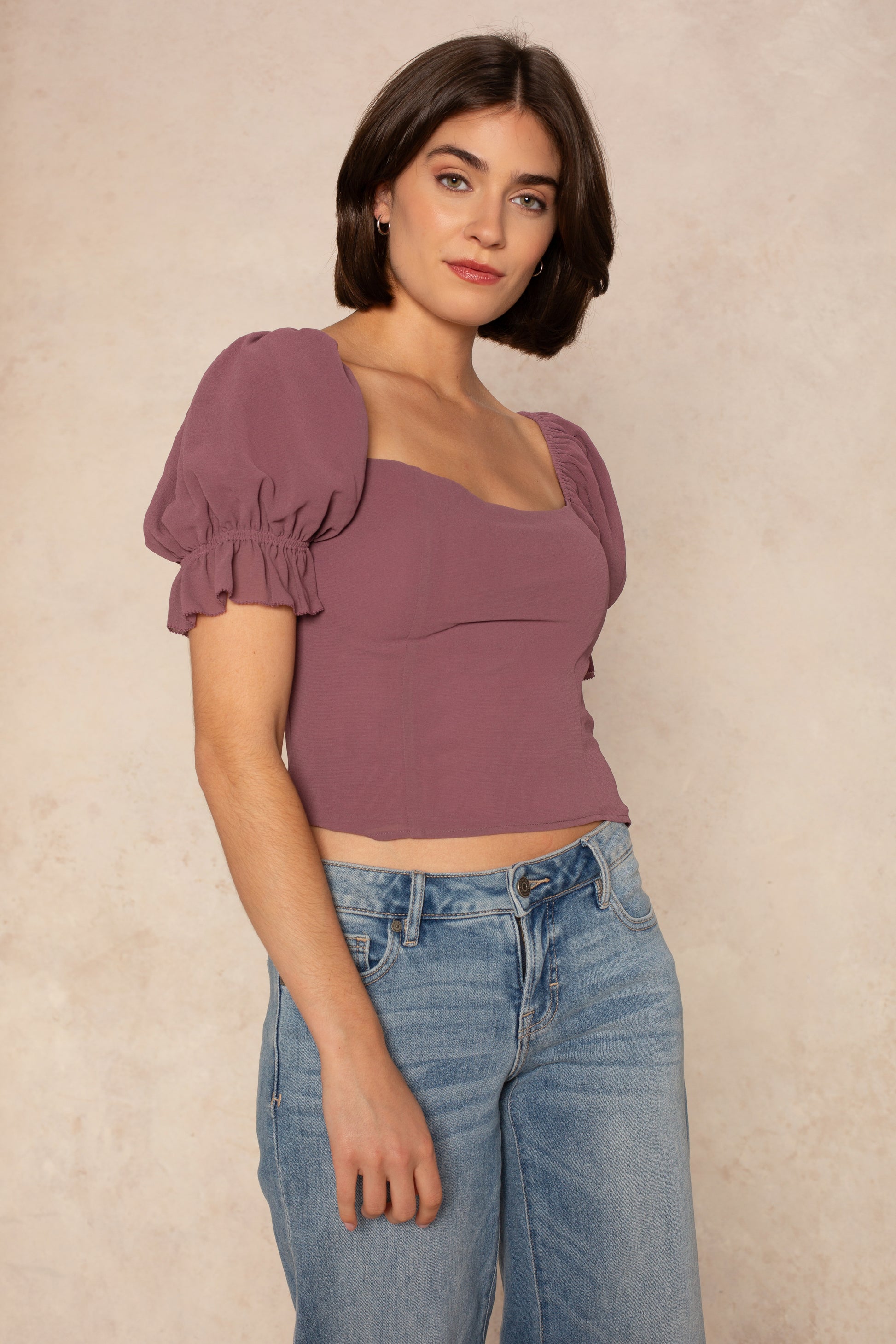 A woman with short brown hair wears the Georgie Hyacinth Top—a mauve puff sleeve blouse—paired with light blue jeans, standing against a neutral textured background and looking at the camera with a slight smile.