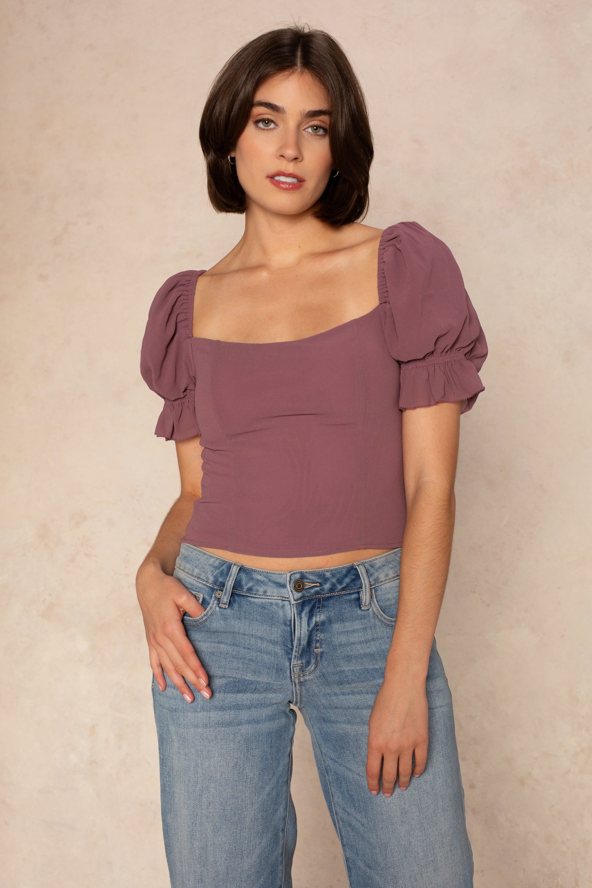 A woman with short brown hair wears the Georgie Hyacinth Top in purple, featuring puff sleeves and a square neckline, paired with blue jeans. She stands against a beige background, facing the camera with one hand in her pocket.