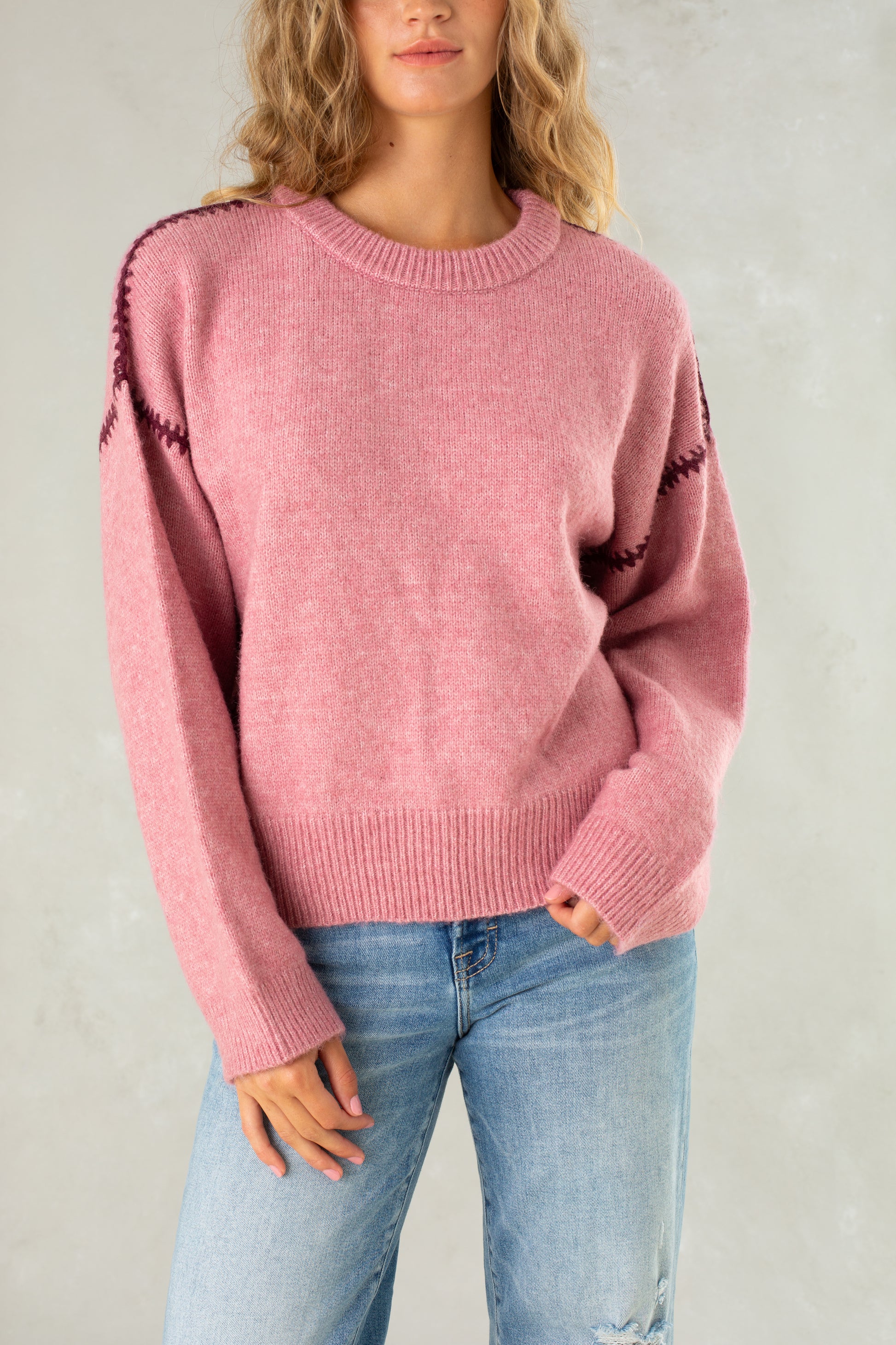A woman with wavy blonde hair models the Isla Jenny Sweater in pink, featuring decorative shoulder stitching, styled with light blue jeans. She stands against a light gray background, one hand in her pocket and her face partially visible.