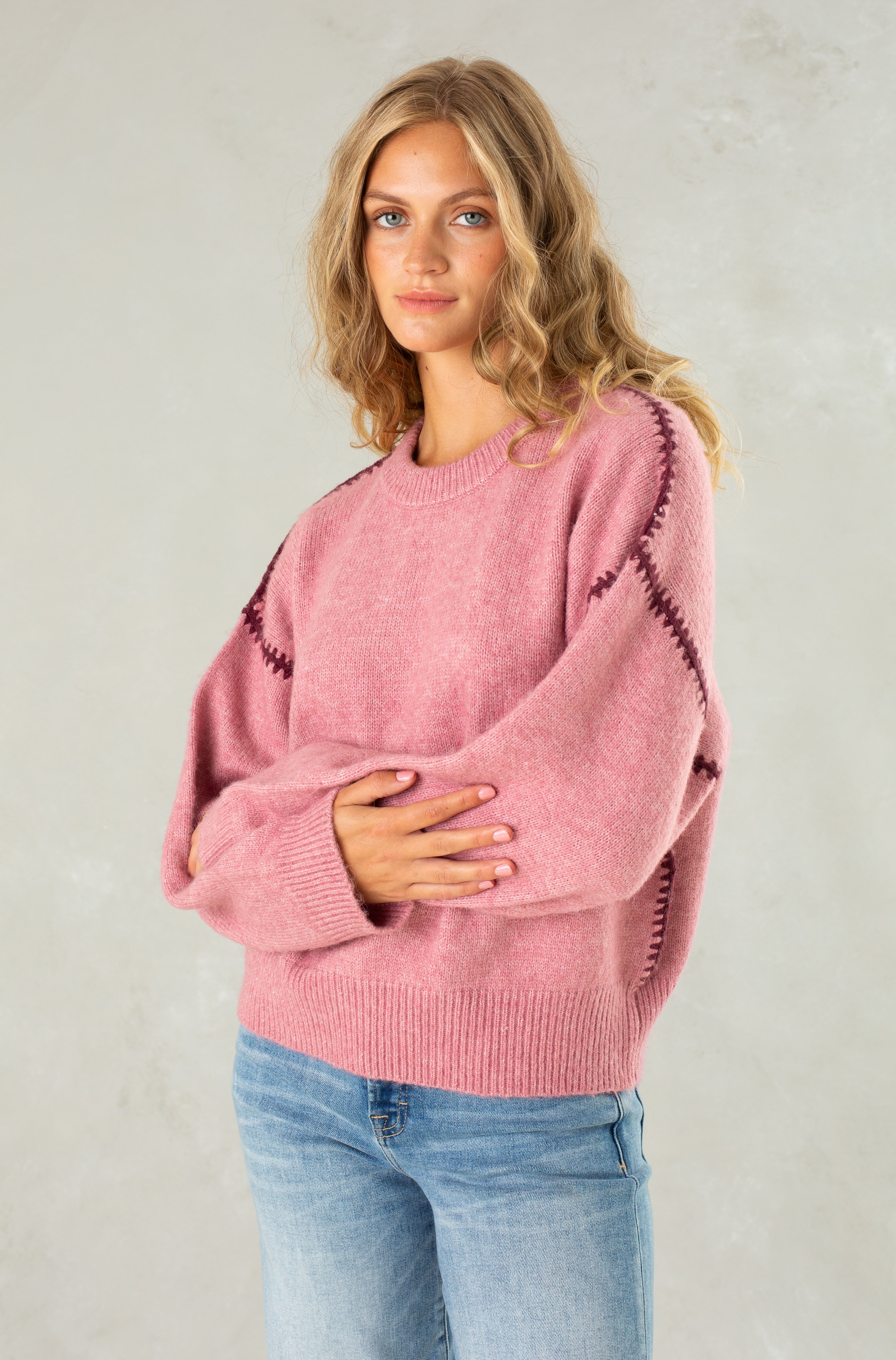 A young woman with wavy blonde hair models Isla's The Jenny Sweater, a relaxed-fit pink crew neck with dark sleeve stitching, paired with blue jeans against a light gray background, gazing at the camera with a relaxed expression.