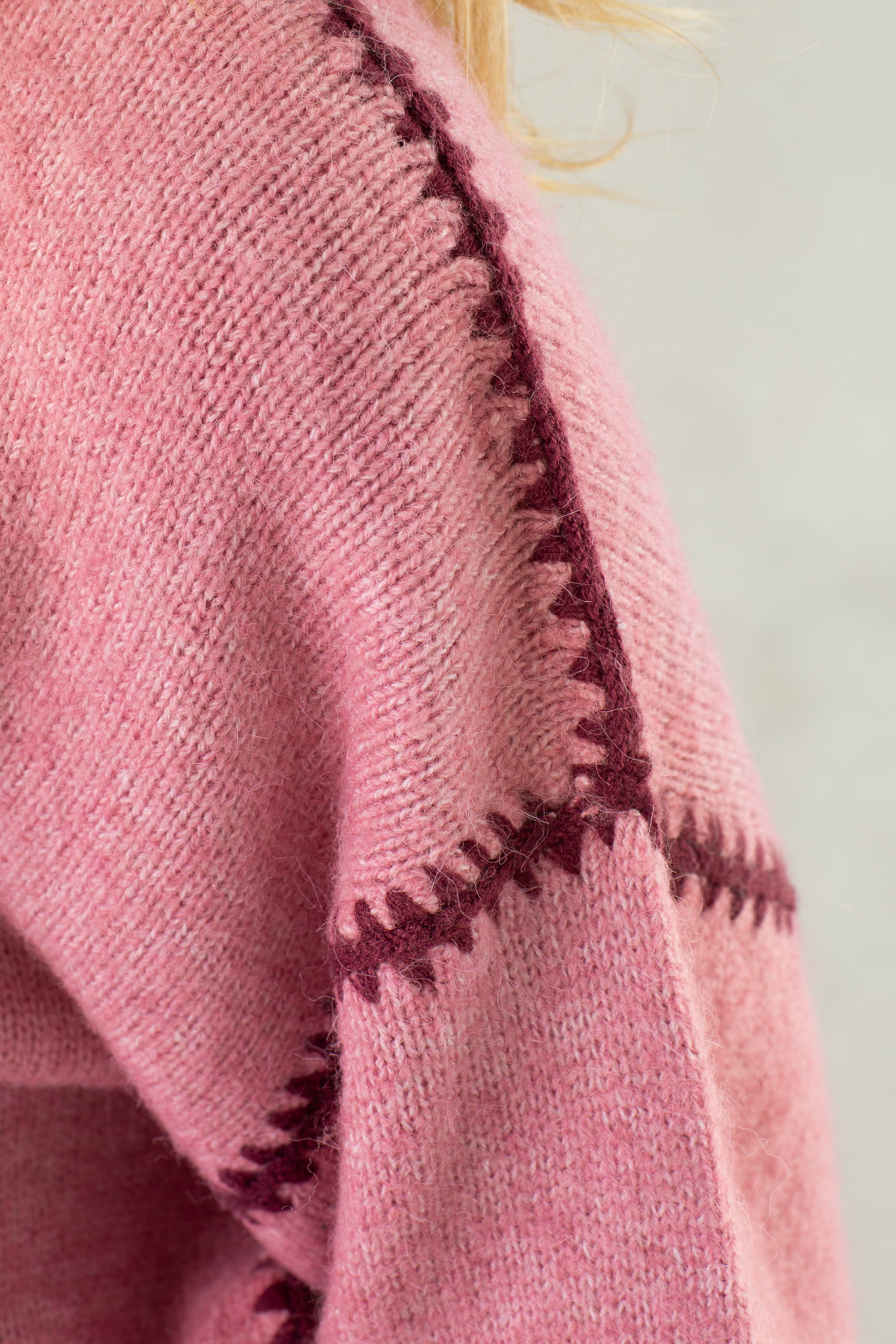 Close-up of a person wearing The Jenny Sweater by Isla, a relaxed-fit crew neck in soft pink with dark maroon zigzag stitching and subtle crochet seam details. Part of the person's blonde hair is visible at the top right.