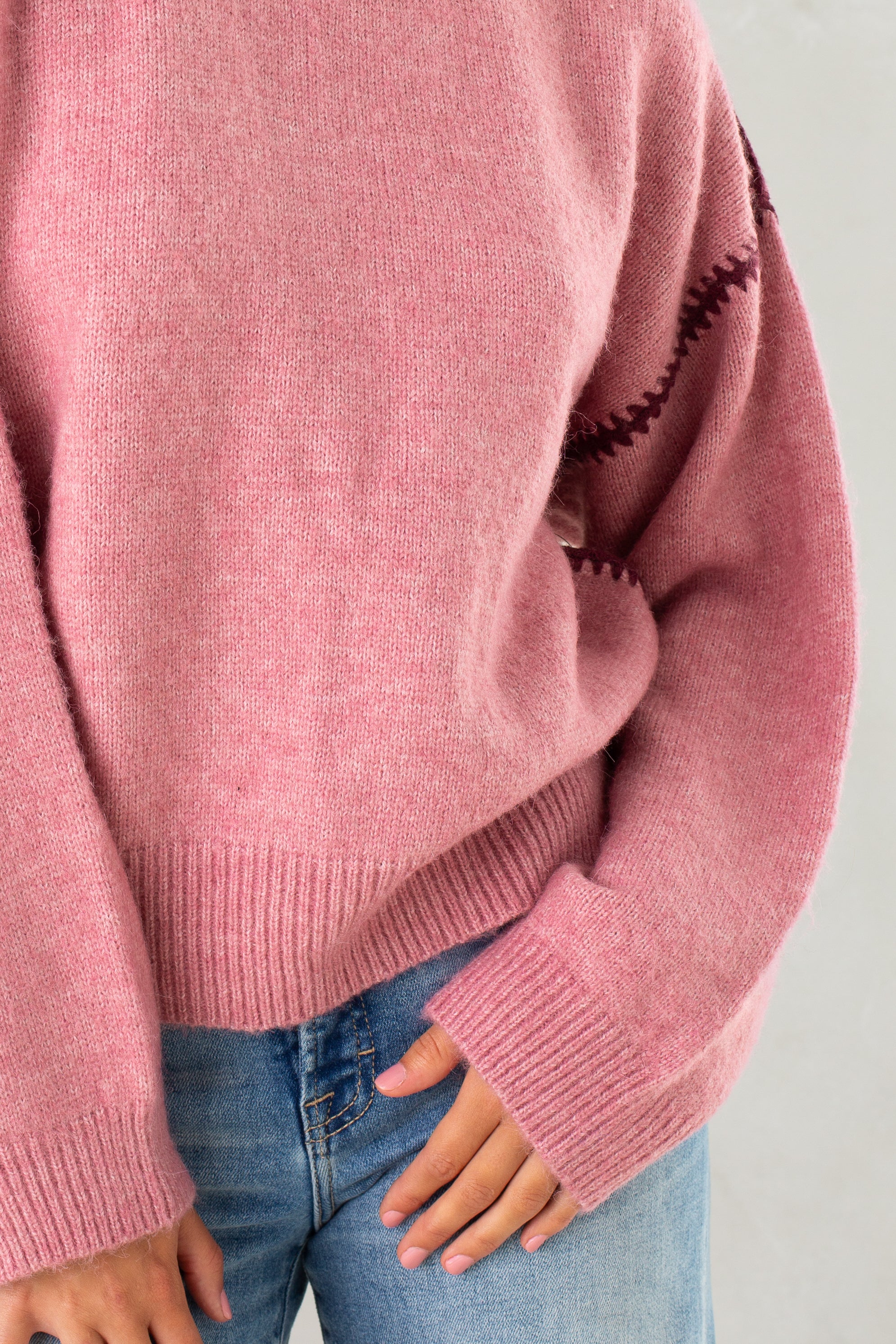 A person wears Isla’s The Jenny Sweater—a pink, relaxed-fit crew neck with subtle crochet sleeve detailing—paired with light blue jeans. Only their torso and hands show, set against a neutral background.