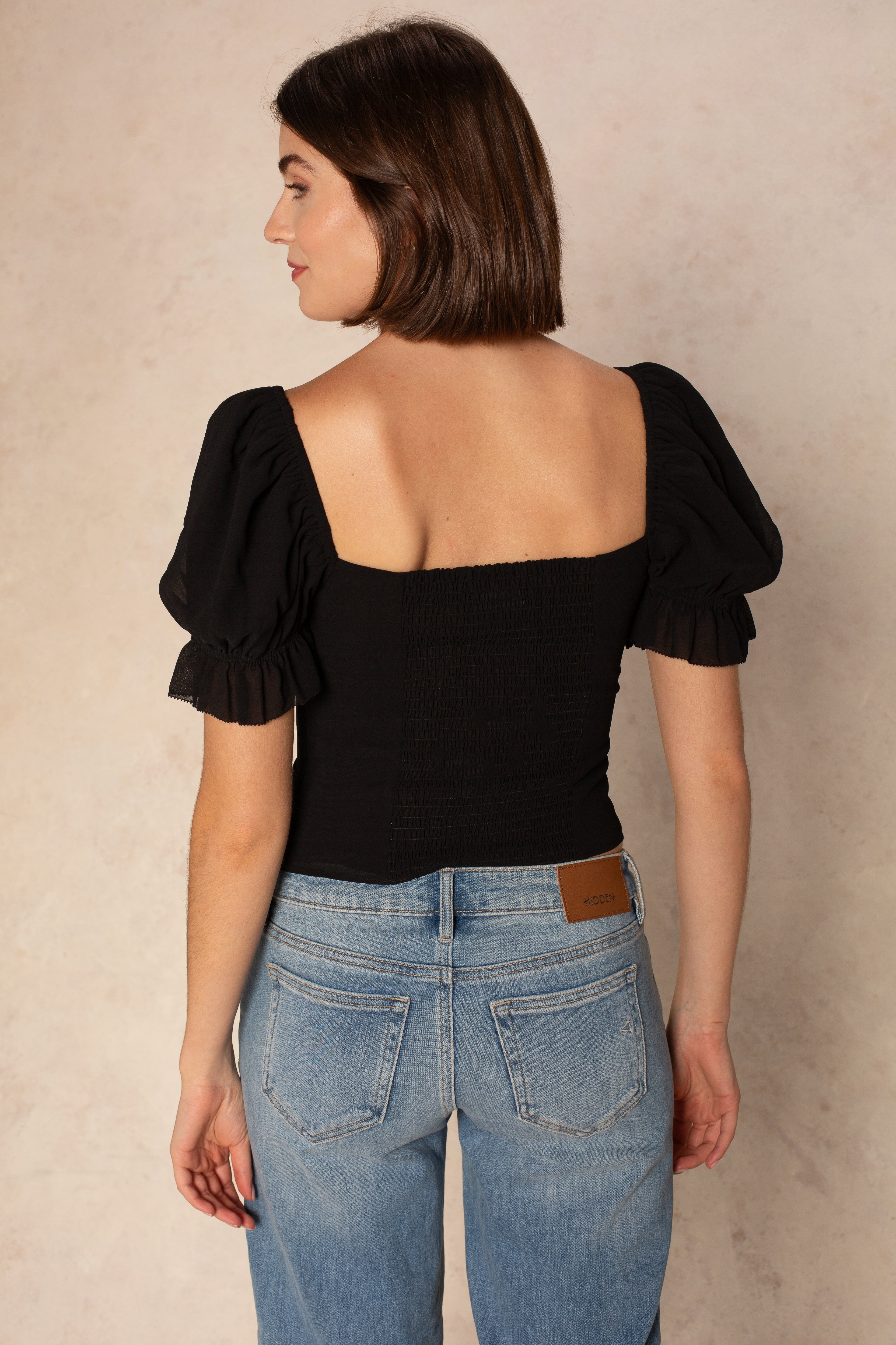 A woman with short brown hair faces away from the camera, showcasing the Georgie Hyacinth Top— a black cropped style with puff sleeves—paired with light blue jeans against a neutral background.