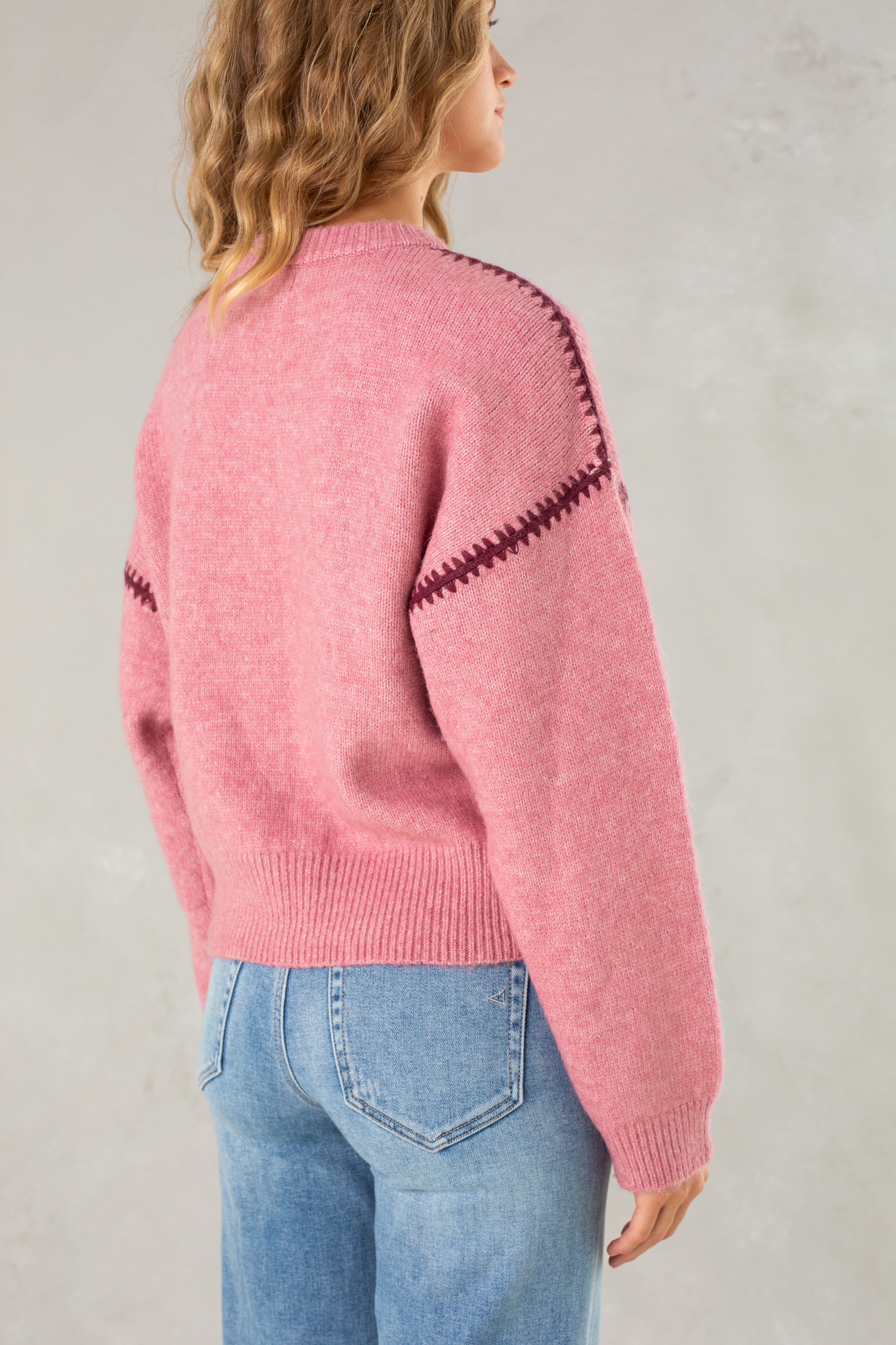 A woman with wavy, shoulder-length hair is seen from the back wearing Isla’s The Jenny Sweater— a pink crew neck with dark zigzag shoulder details— and light blue jeans, posed against a light gray background.