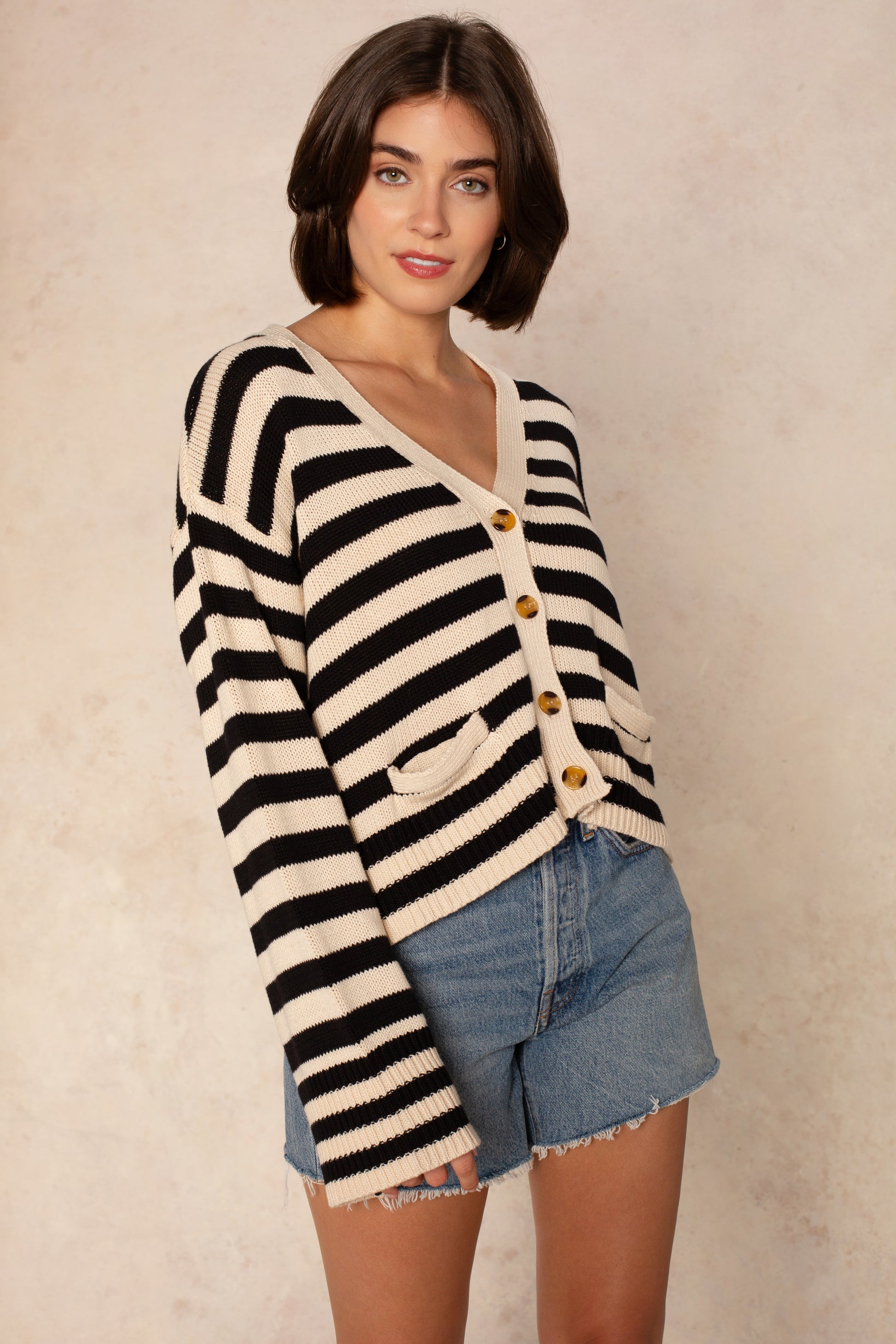A woman with short brown hair wears Isla’s Heather Stripe Crochet Cardigan in Black Multi and denim shorts, standing against a light beige background.
