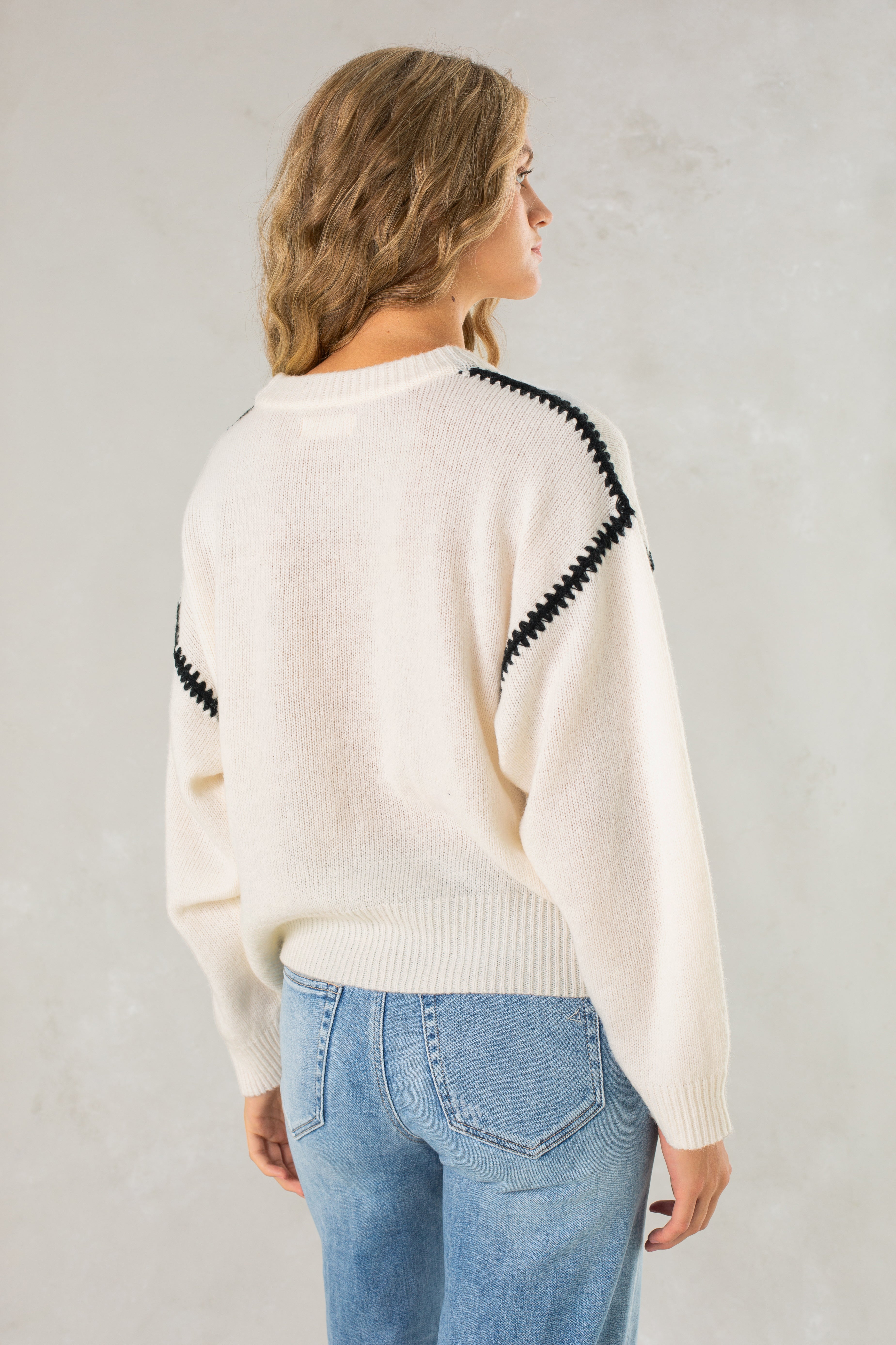 A woman with wavy blonde hair faces away from the camera, wearing Isla's The Jenny Sweater—a relaxed-fit crew neck with black zigzag stripes on the shoulders—and light blue jeans against a light gray background.