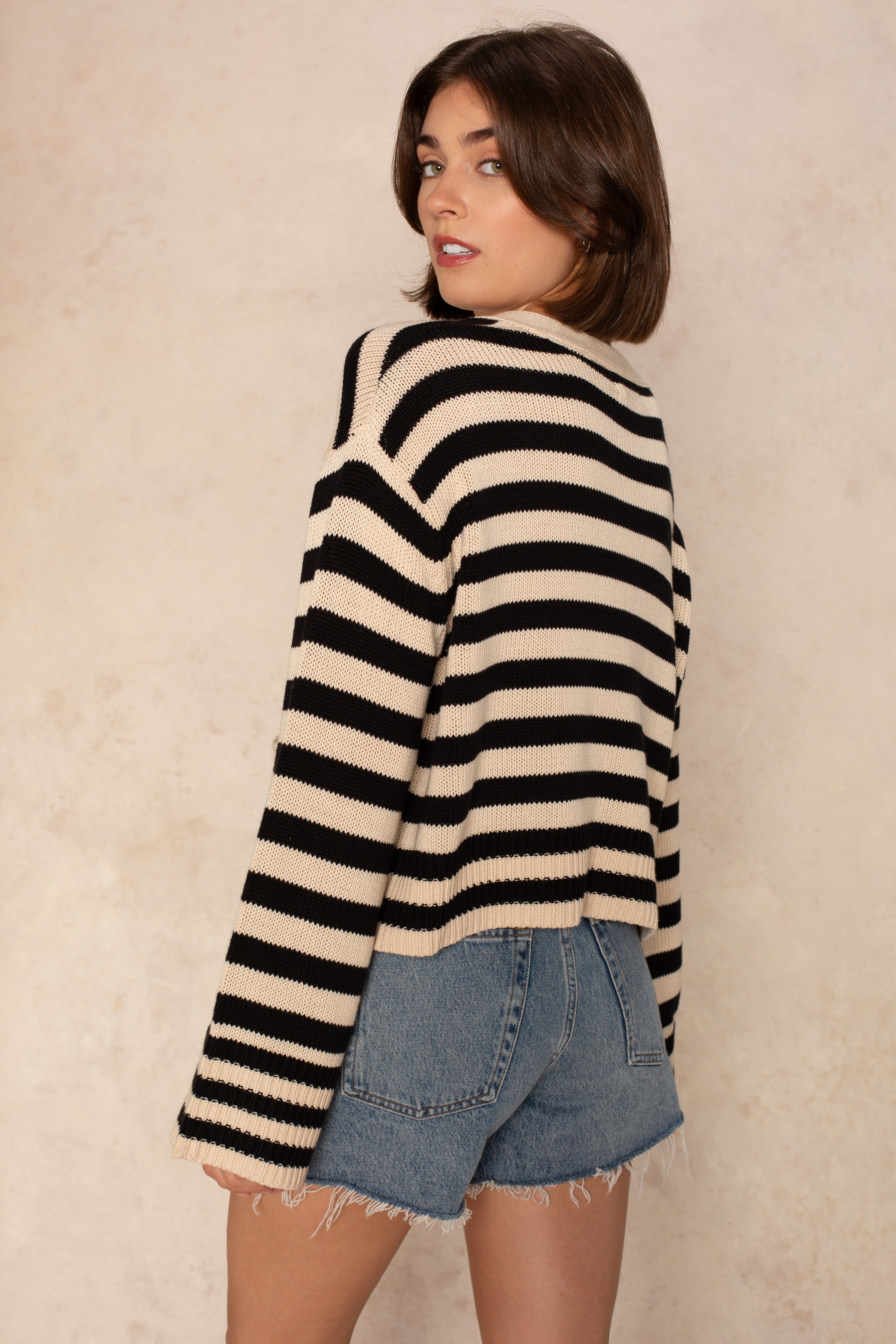 A woman with short brown hair models the Isla Heather Stripe Crochet Cardigan, styled oversized and cropped, with denim shorts. She stands sideways, looking over her shoulder against a light, neutral background.