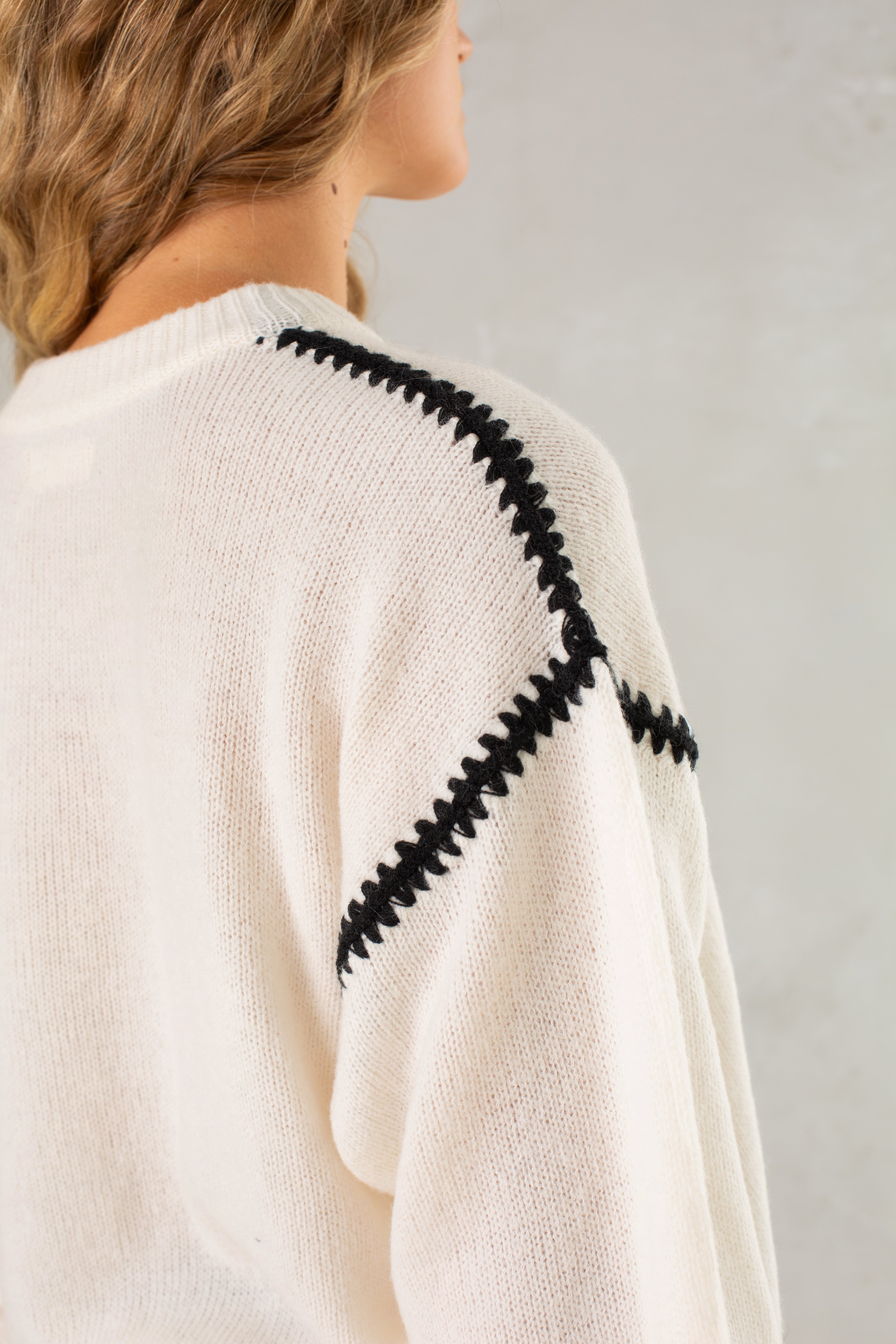 A close-up of Isla's The Jenny Sweater, a relaxed-fit cream crew neck with black zigzag stitching on the shoulder and sleeve seams. Wavy hair partly covers the back, set against a plain light background.