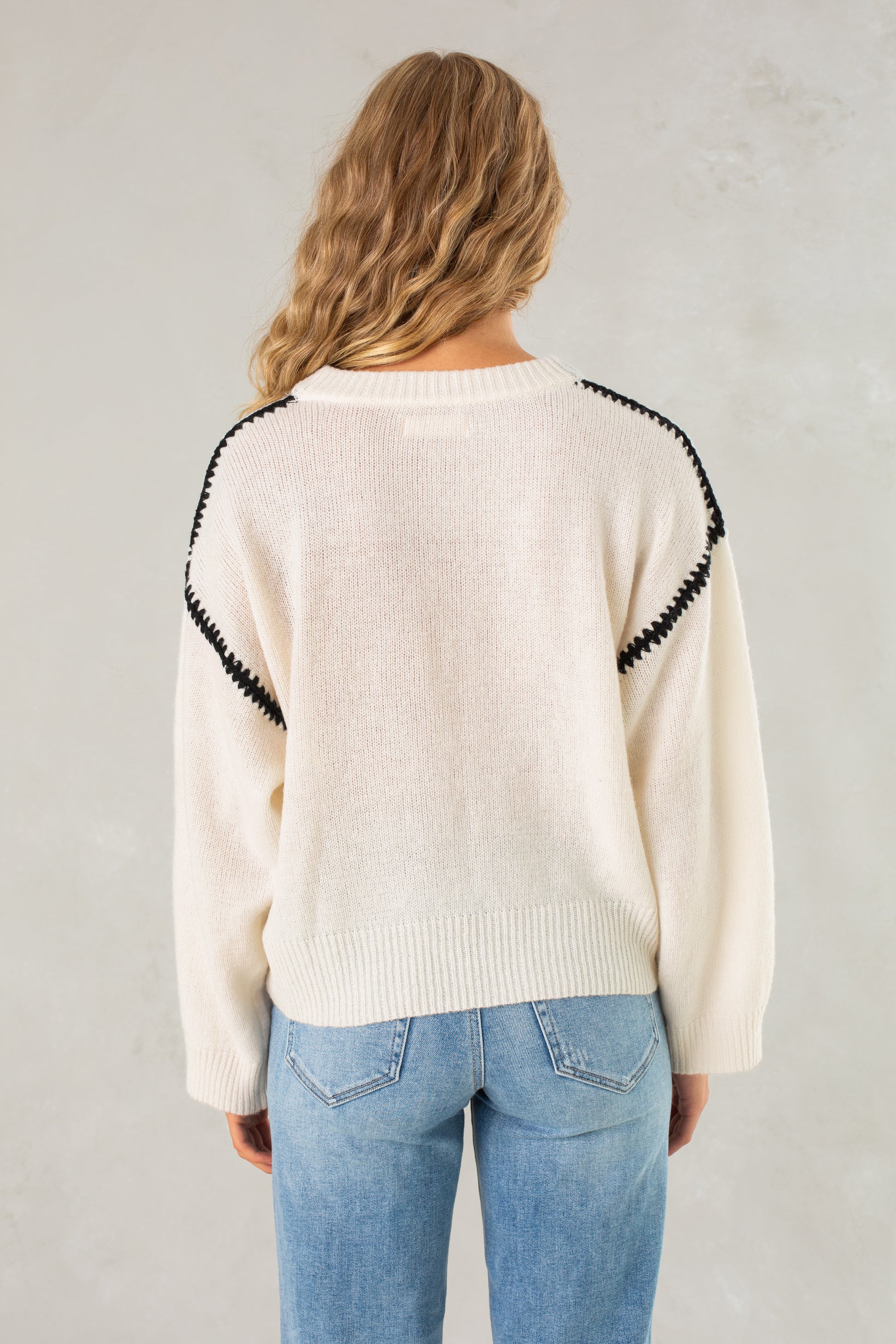 A person with long wavy blonde hair wears Isla's The Jenny Sweater, a relaxed-fit crewneck with black shoulder detailing, and blue jeans, standing with their back to a light gray textured wall.