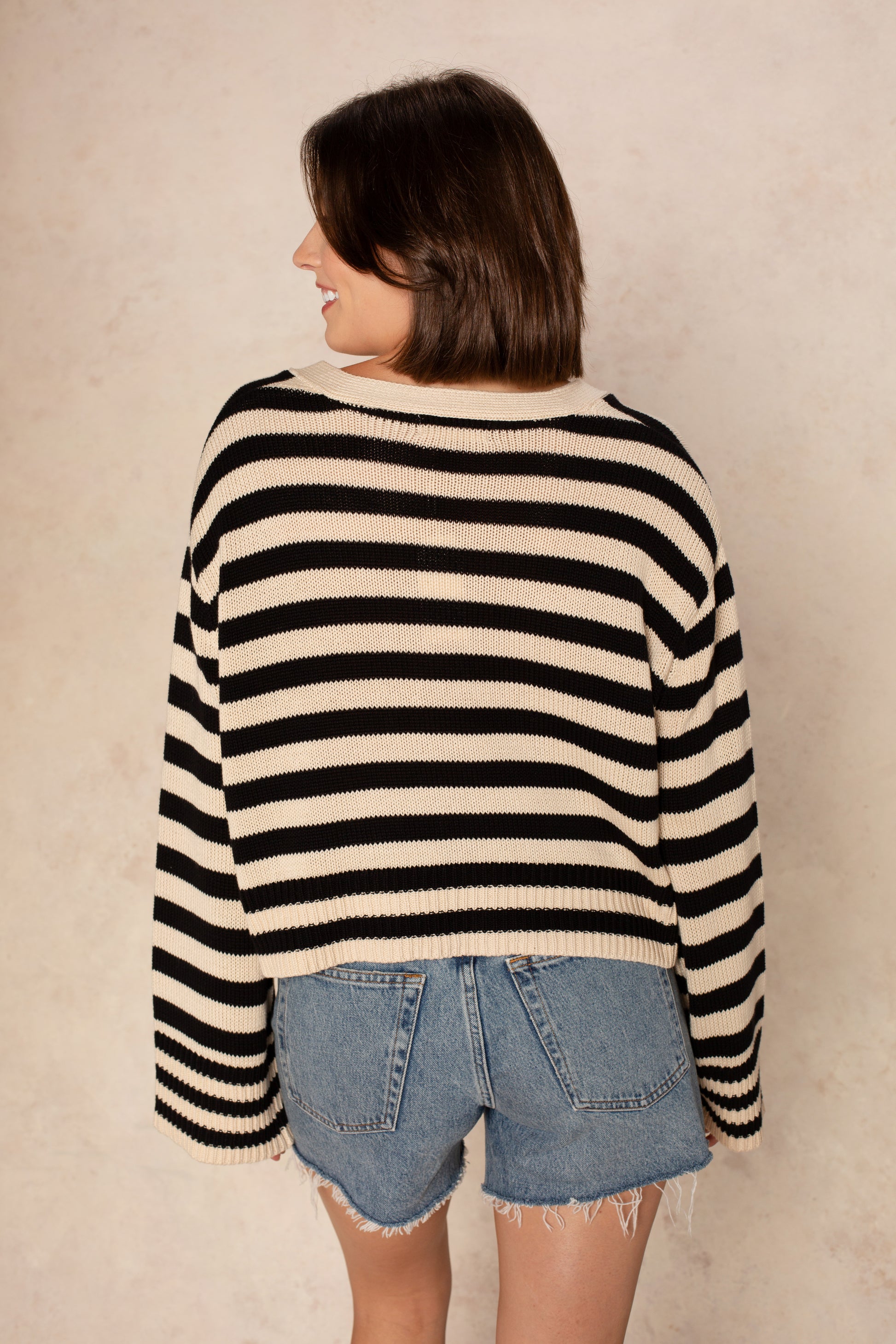 A woman with short brown hair smiles as she faces away, wearing the Isla Heather Stripe Crochet Cardigan in blue and white stripes with frayed denim shorts, standing against a neutral beige background.