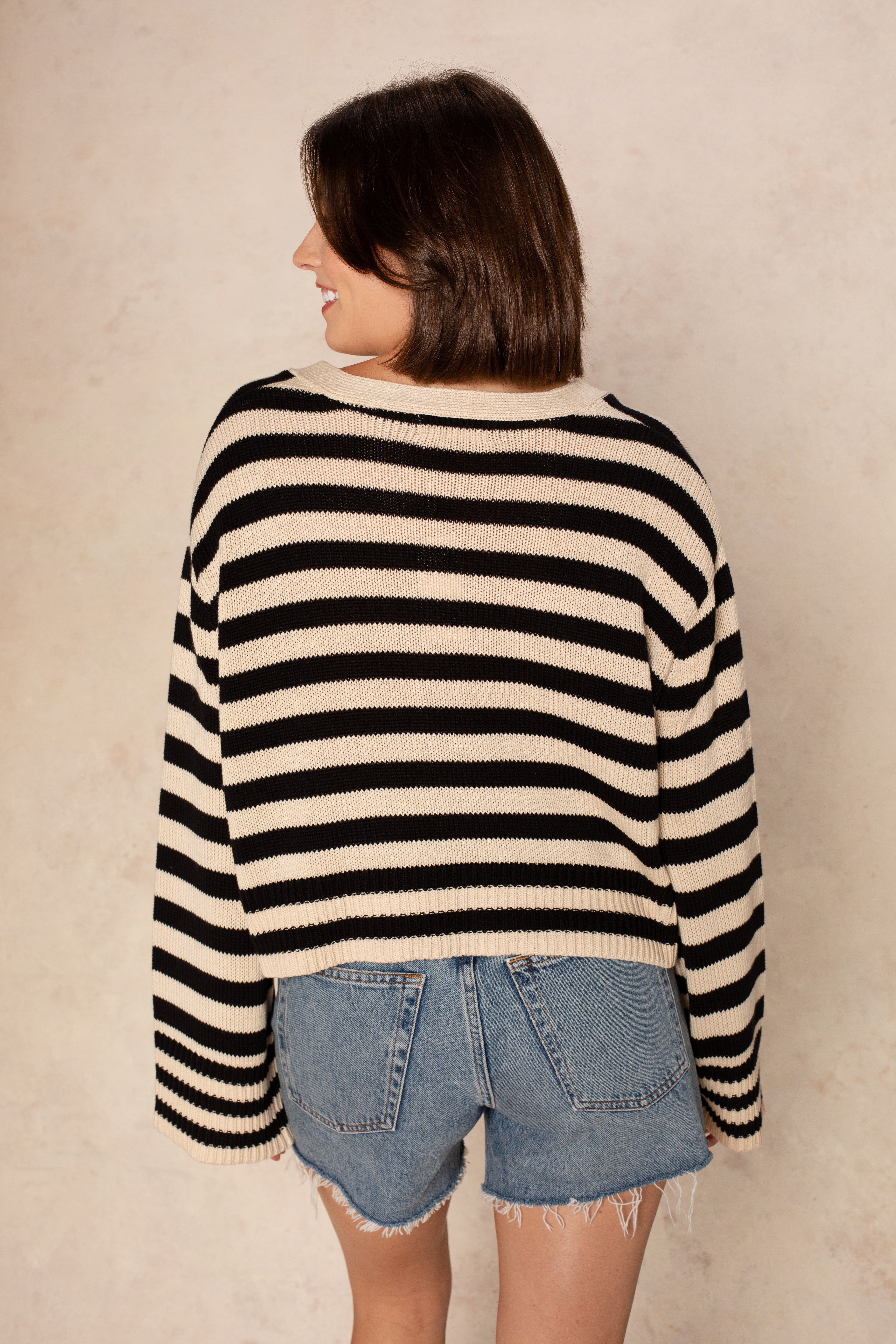 A woman with short brown hair smiles as she faces away, wearing the Isla Heather Stripe Crochet Cardigan in blue and white stripes with frayed denim shorts, standing against a neutral beige background.