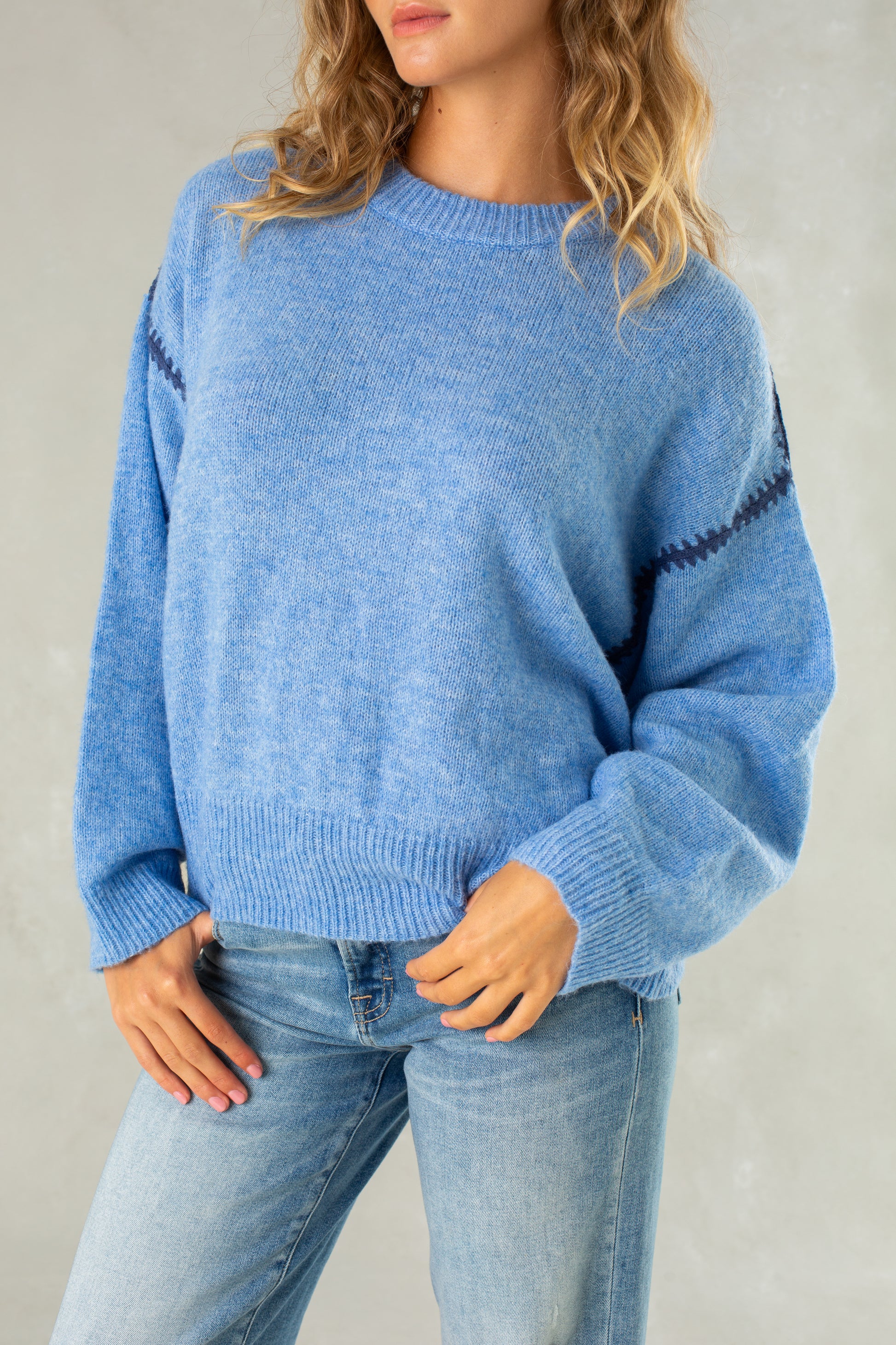 A woman with wavy blonde hair wears Isla’s The Jenny Sweater—a relaxed-fit, light blue pullover with dark blue zigzag sleeve stripes—paired with light blue jeans, standing against a plain light background.