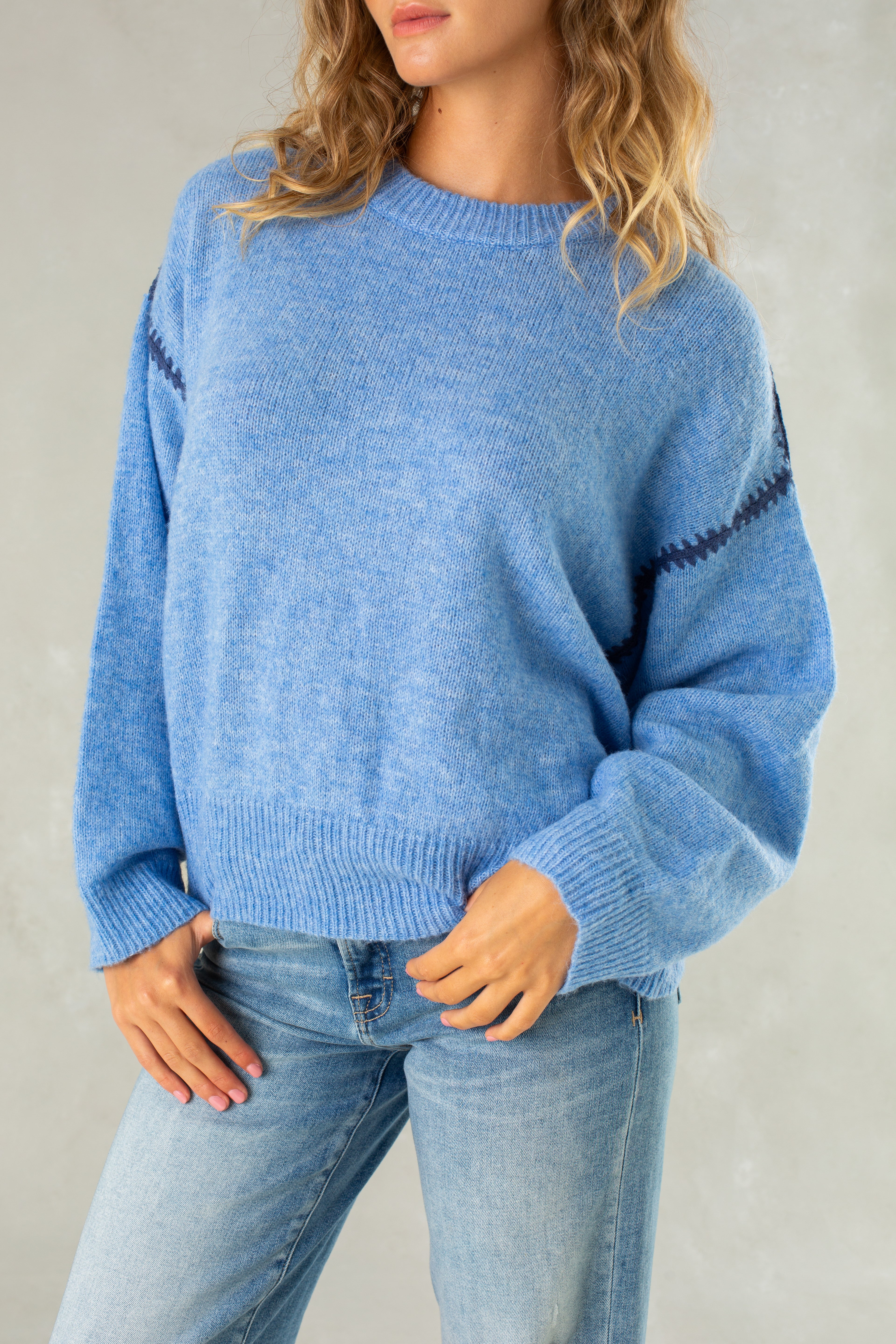 A woman with wavy blonde hair wears Isla’s The Jenny Sweater—a relaxed-fit, light blue pullover with dark blue zigzag sleeve stripes—paired with light blue jeans, standing against a plain light background.