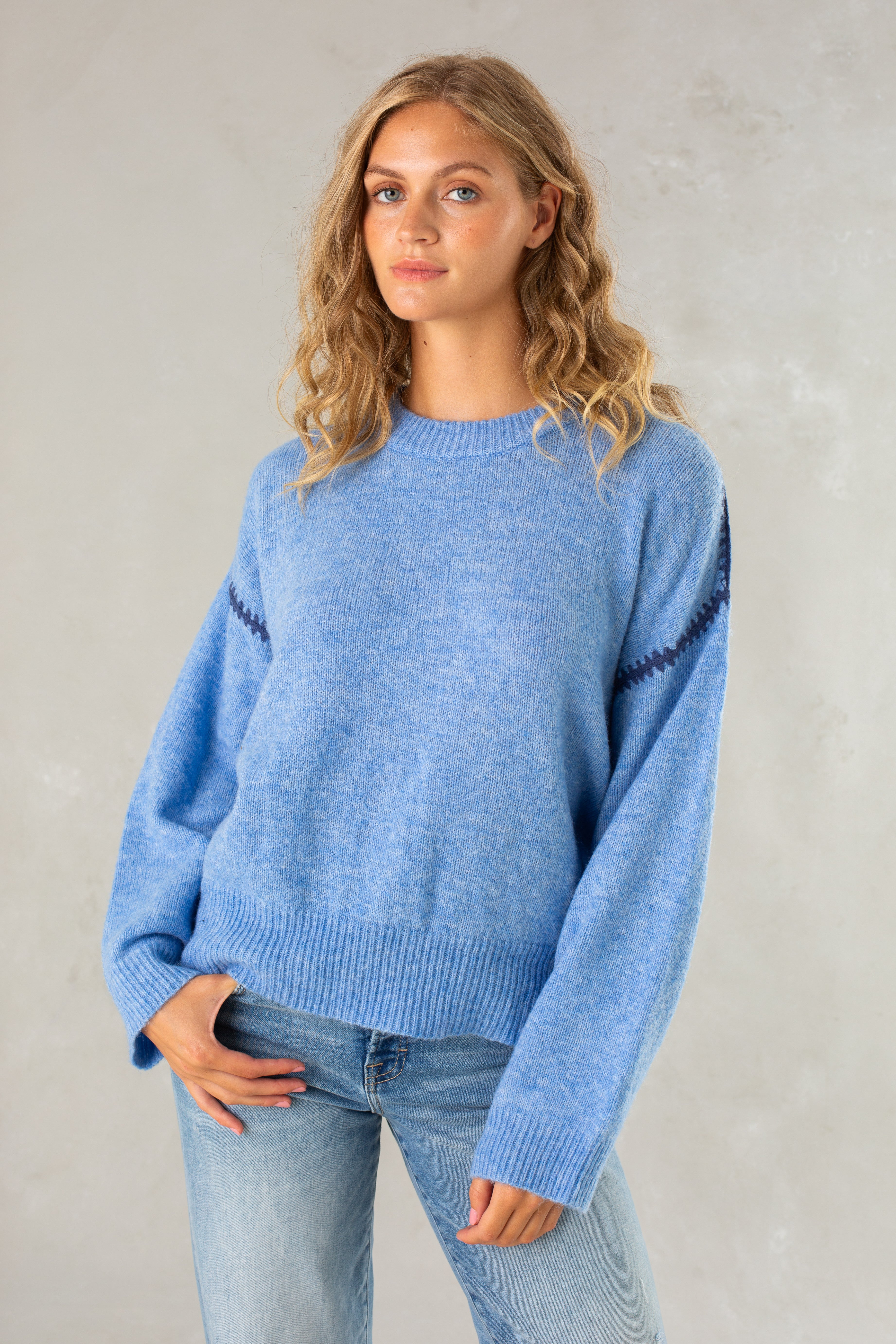 A young woman with wavy blonde hair wears the Isla Jenny Sweater in blue paired with light blue jeans, standing against a plain, light-colored background.