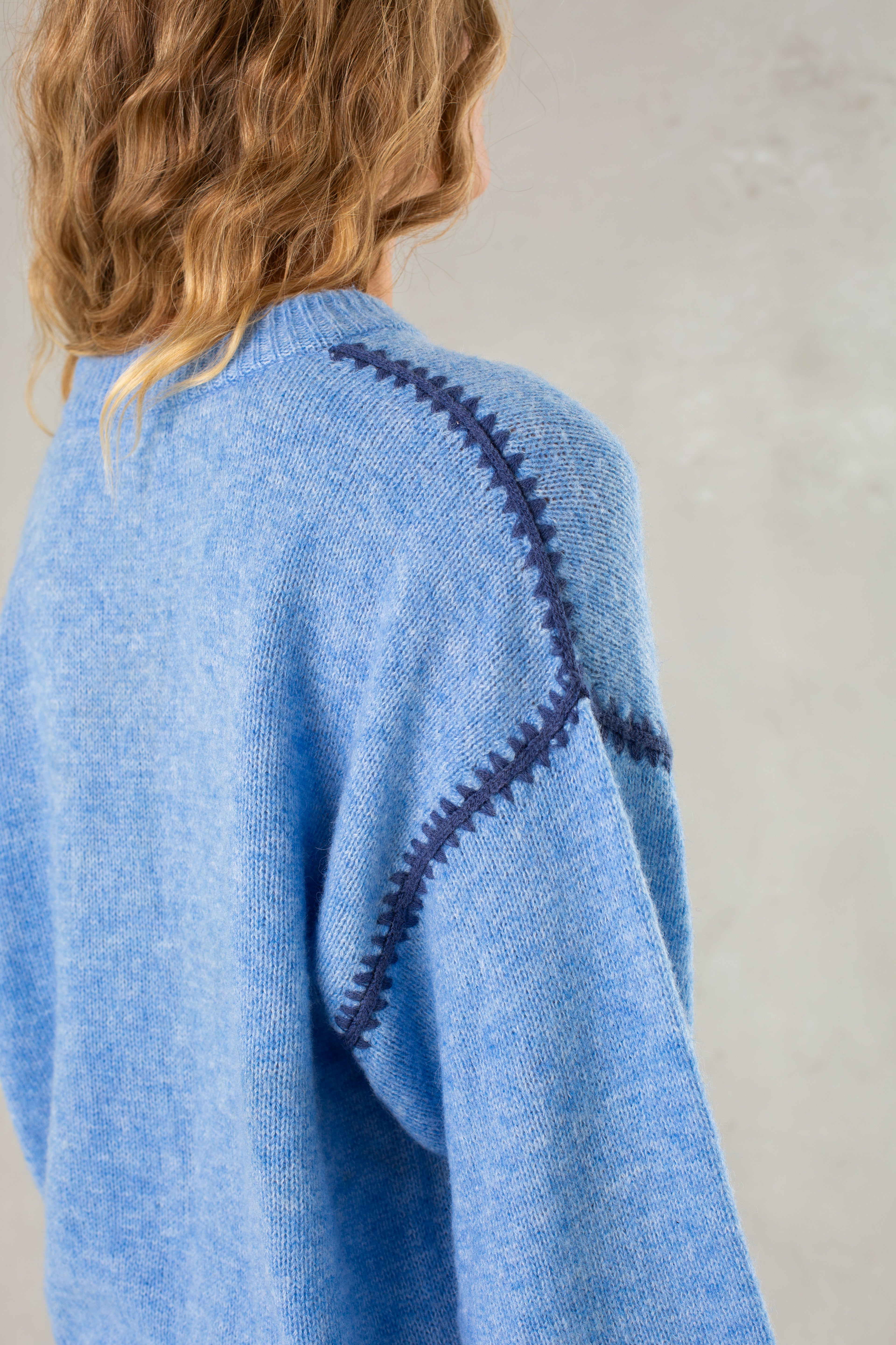 A person with wavy, light brown hair wears Isla's The Jenny Sweater, a relaxed-fit light blue pullover with dark blue decorative stitching on the shoulders and sleeves, standing against a plain light background.