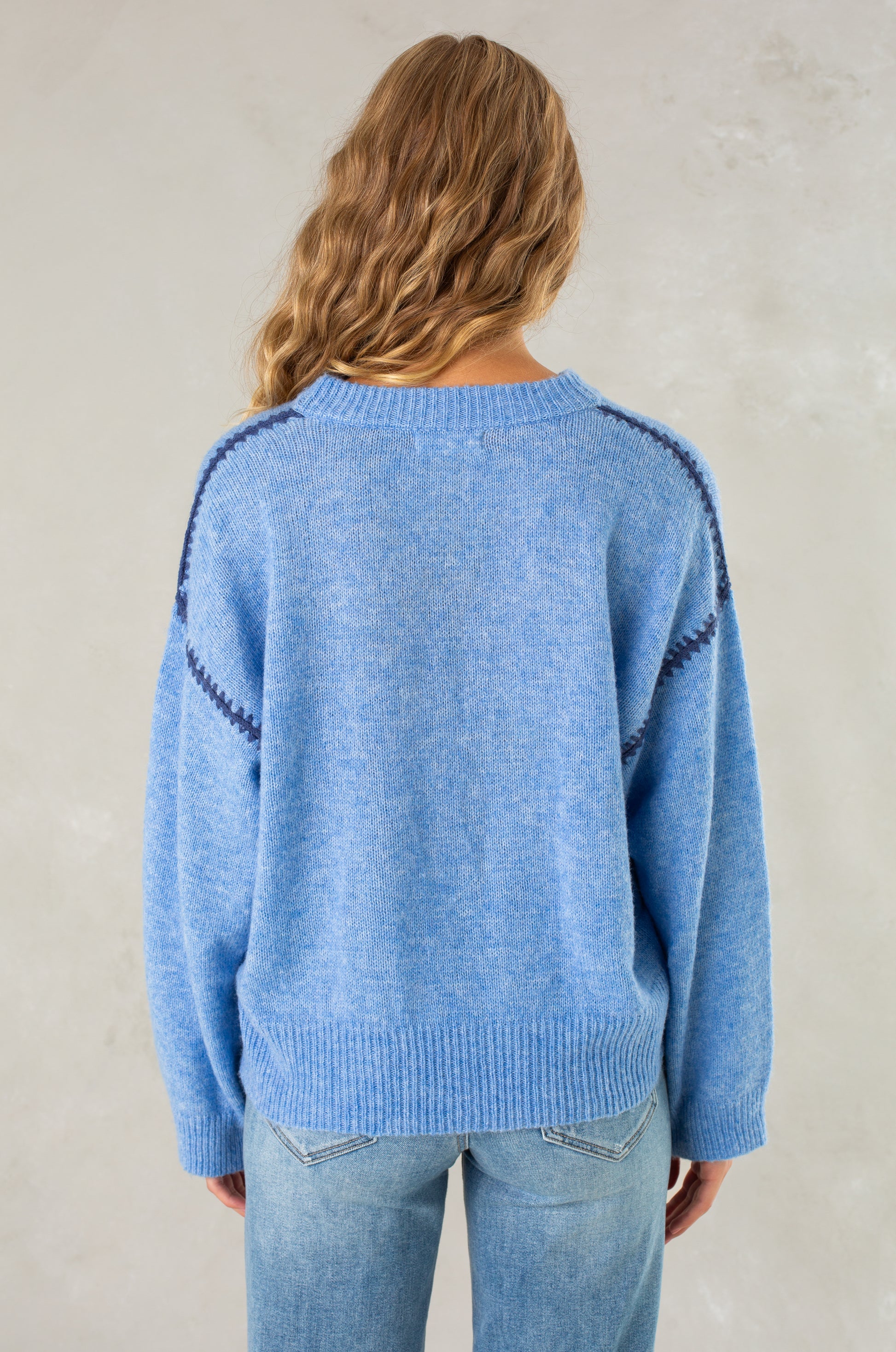 A person with long wavy hair models Isla’s The Jenny Sweater, a relaxed-fit light blue crew neck with dark blue sleeve accents, paired with light blue jeans, standing back to the camera against a neutral background.
