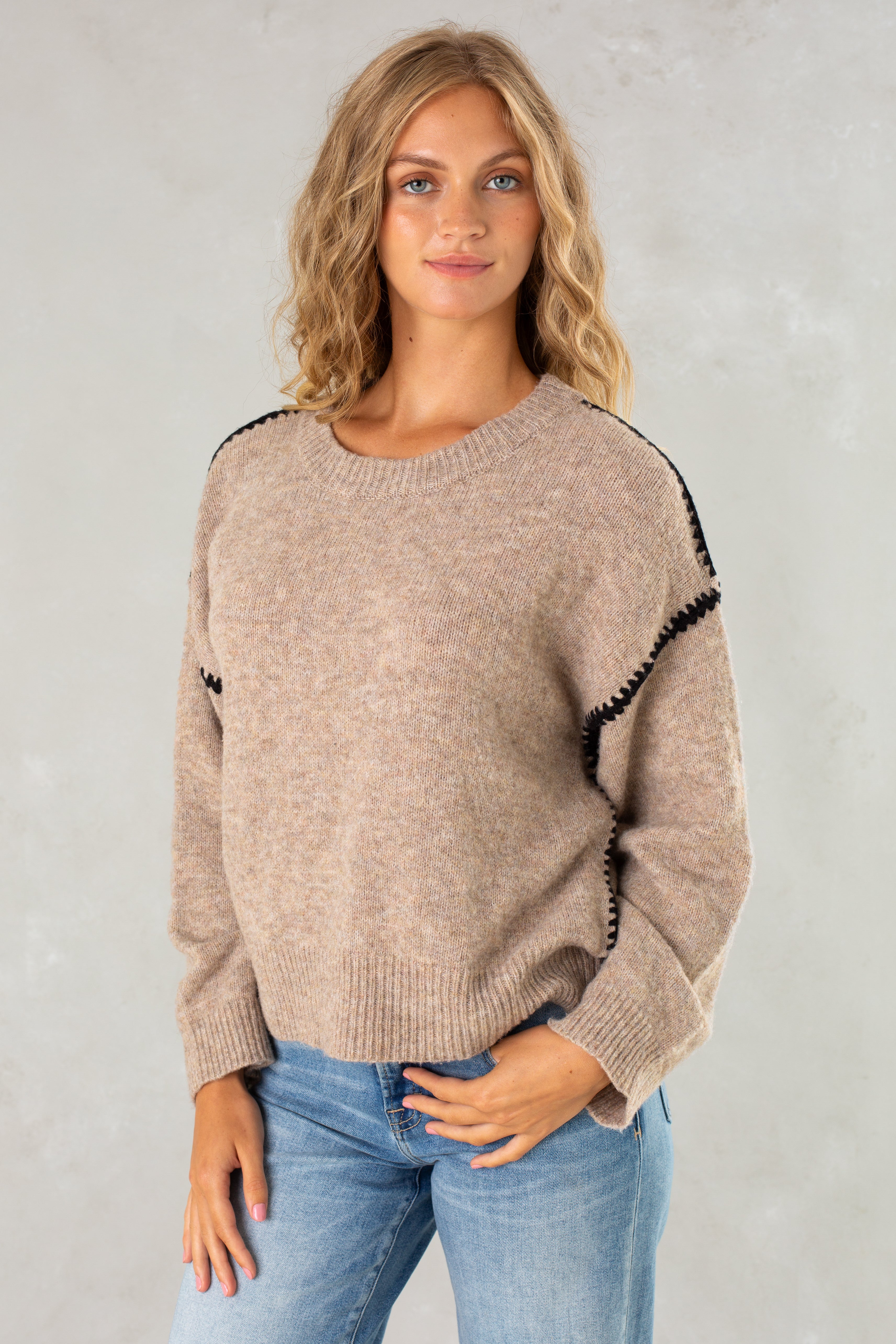 A young woman with wavy blonde hair wears The Jenny Sweater by Isla—a cozy crew neck with black trim—paired with blue jeans, standing against a light gray background and smiling slightly at the camera.