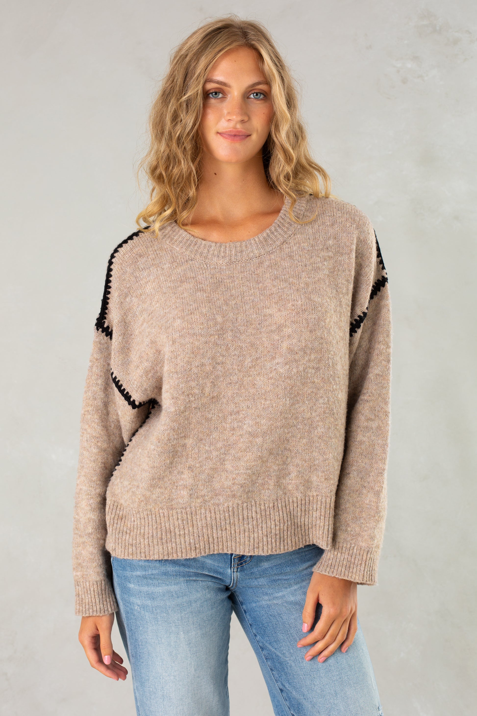 A woman with long, wavy blonde hair models Isla’s The Jenny Sweater in tan—a relaxed-fit pullover with black shoulder trim—paired with light blue jeans, standing against a light gray background and smiling slightly at the camera.