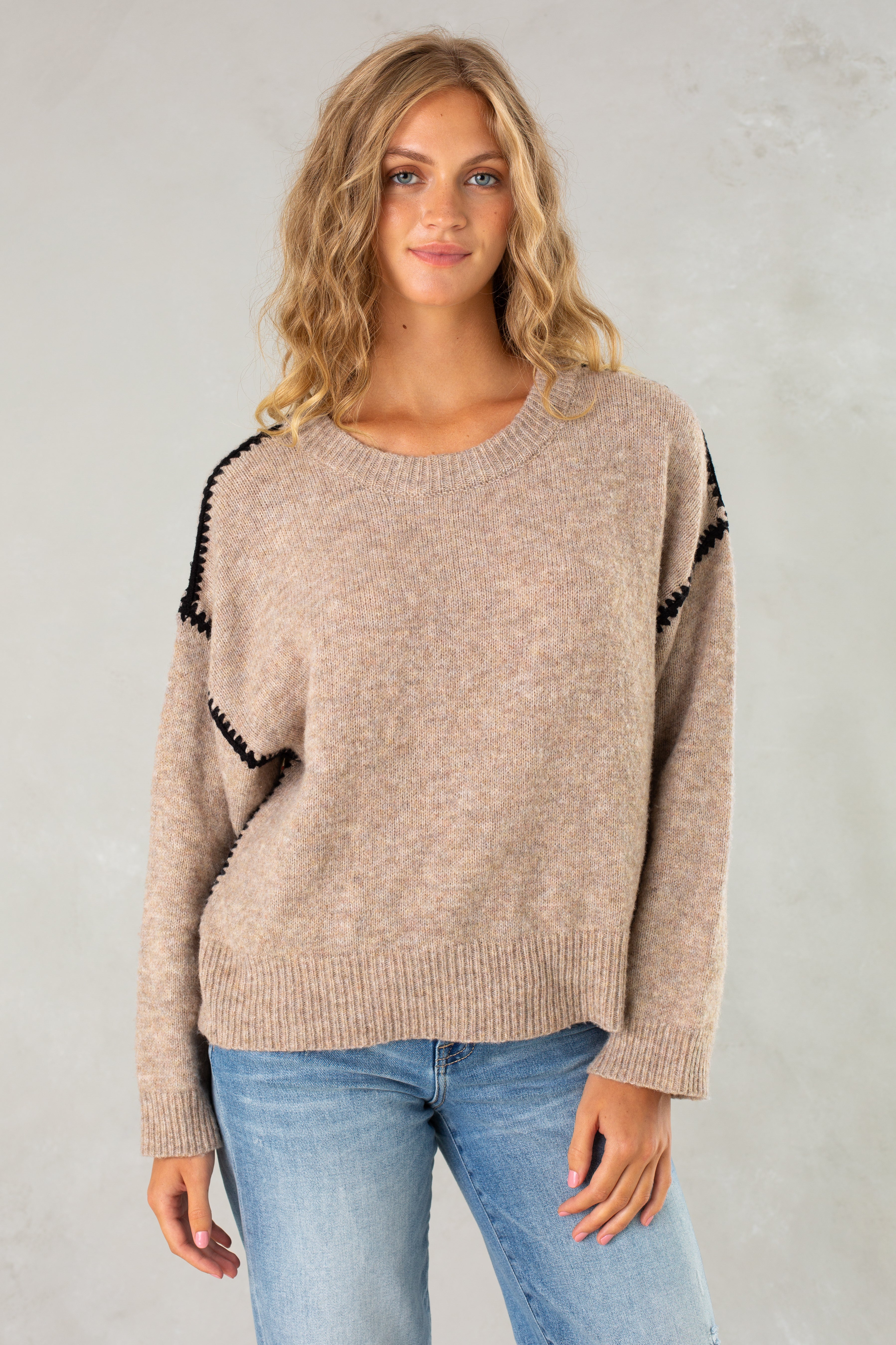 A woman with long, wavy blonde hair models Isla’s The Jenny Sweater in tan—a relaxed-fit pullover with black shoulder trim—paired with light blue jeans, standing against a light gray background and smiling slightly at the camera.
