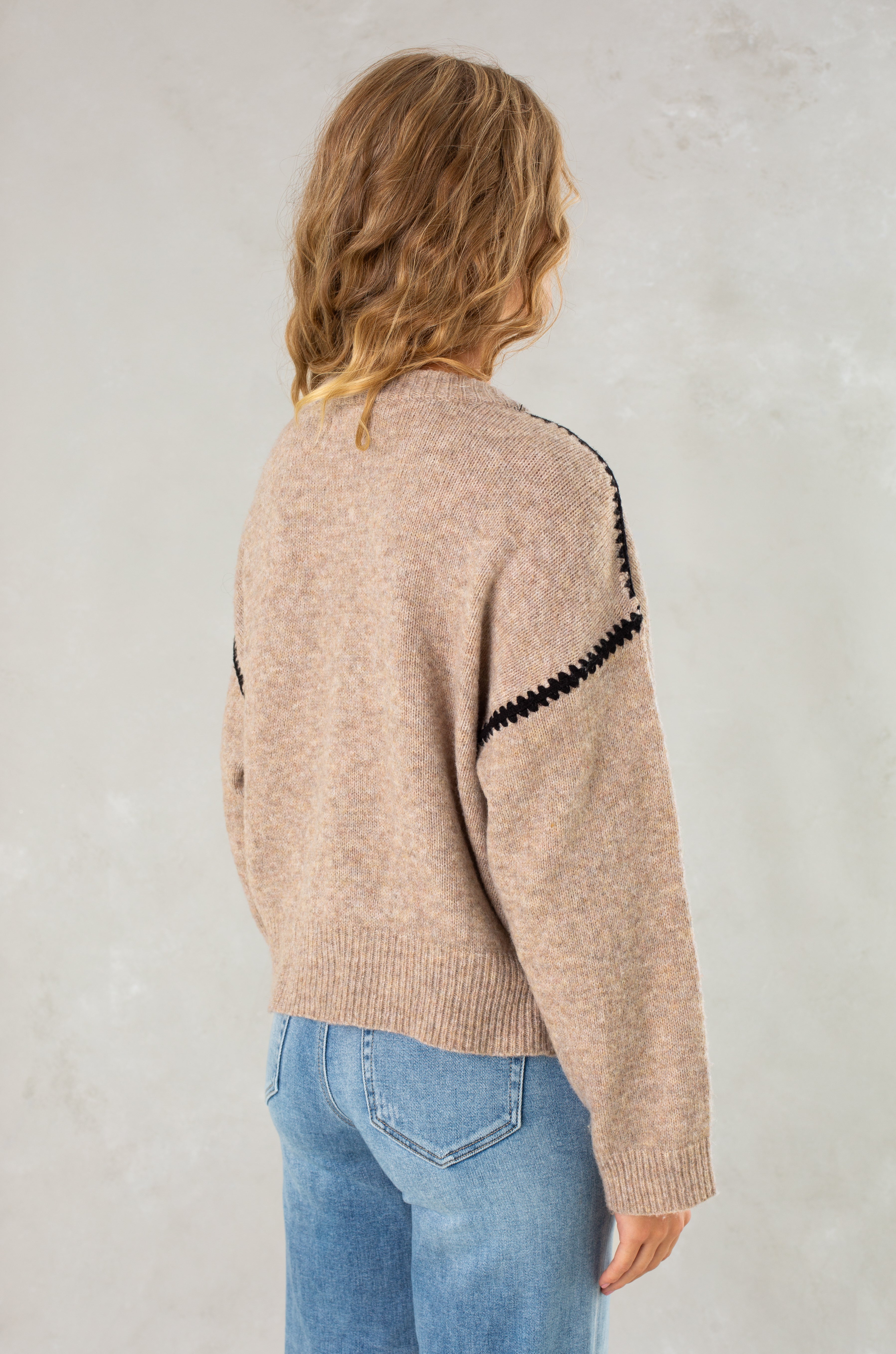 A person with wavy, shoulder-length hair is seen from behind wearing Isla’s The Jenny Sweater, a beige cozy crew neck with black shoulder detailing, paired with blue jeans and standing against a light textured background.