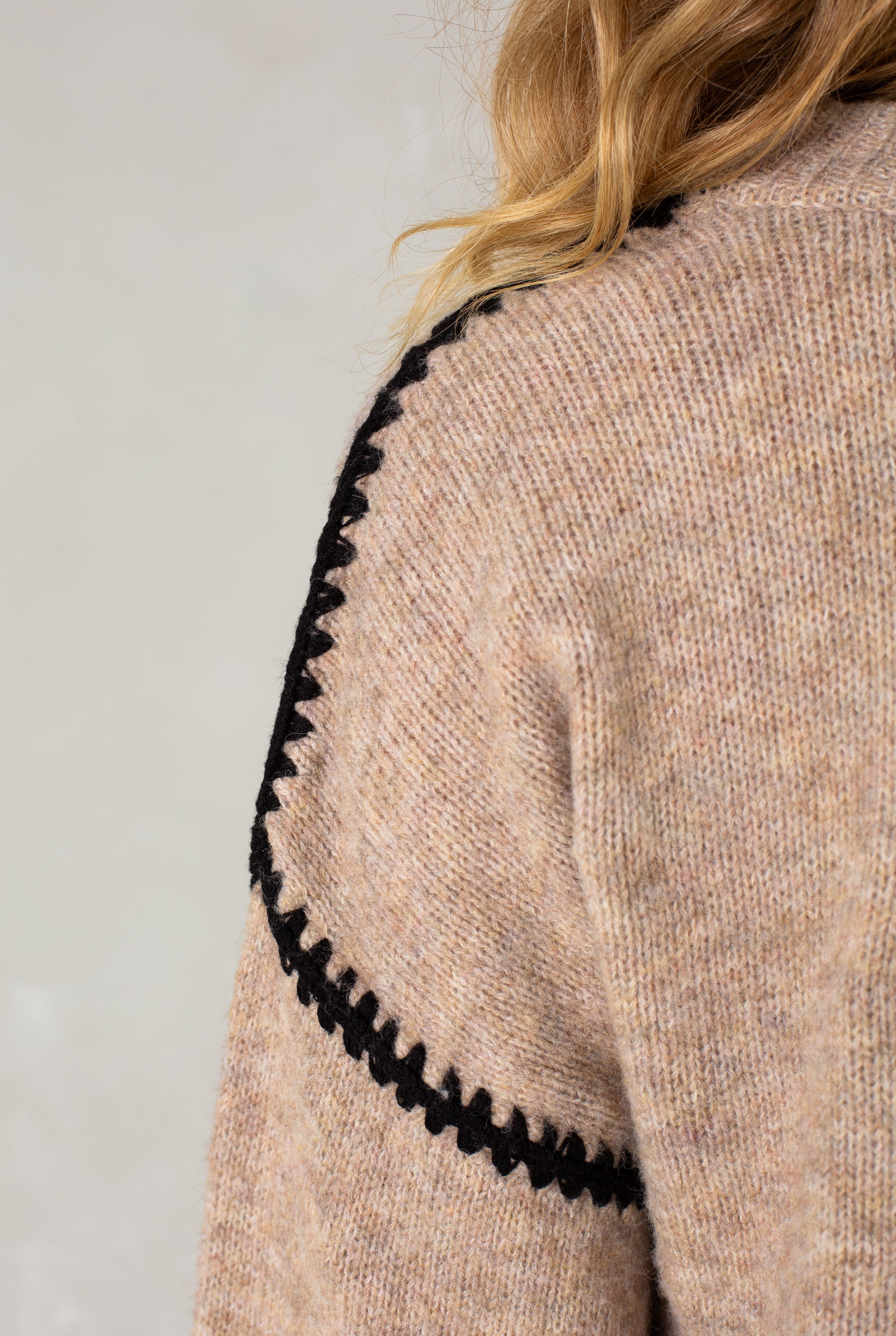 Close-up of a shoulder wearing The Jenny Sweater by Isla, a cozy beige crew neck with black zigzag stitching along the shoulder and arm seam. Light-colored background.