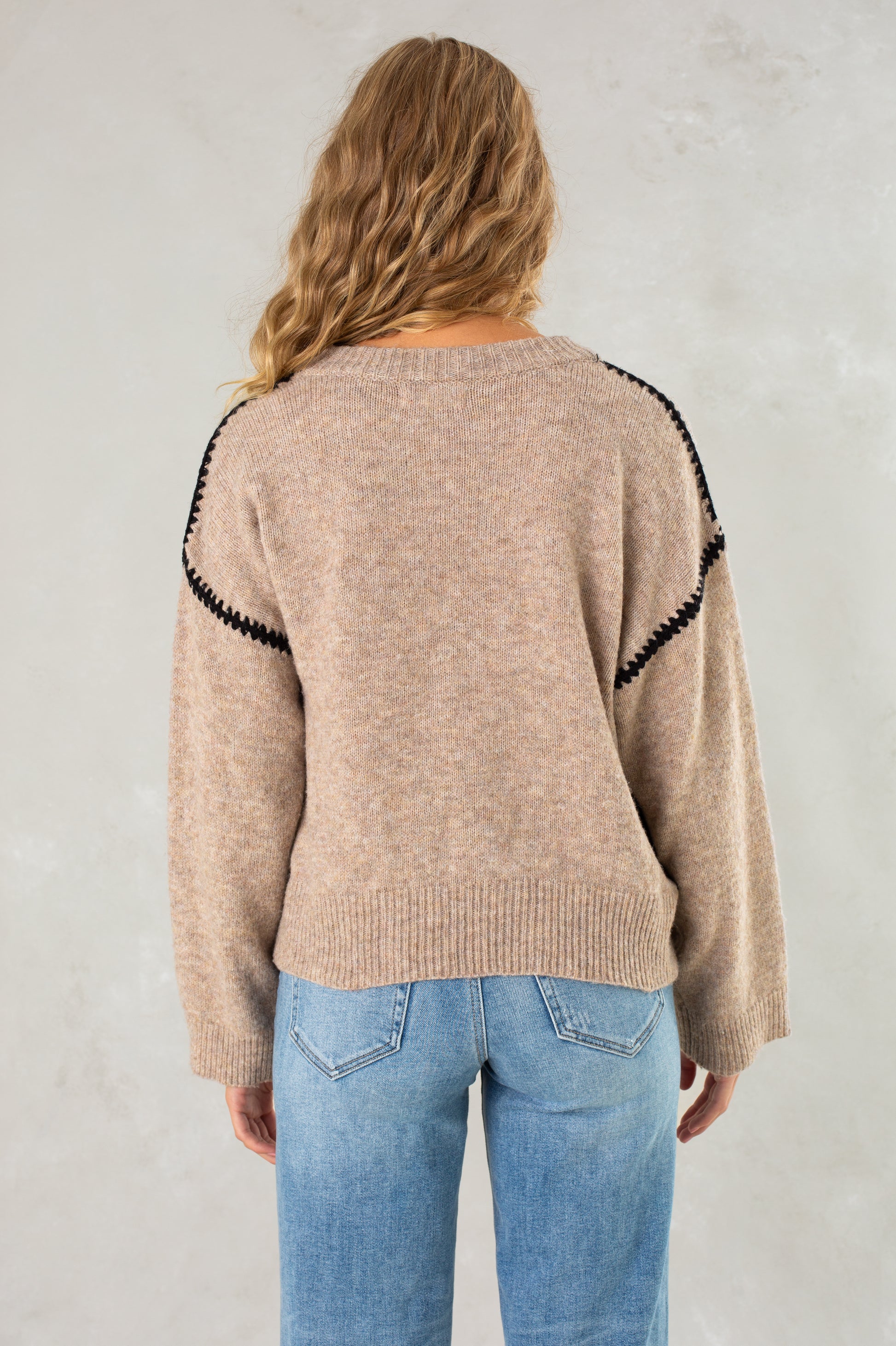 A person with long wavy blonde hair wears Isla's The Jenny Sweater, a cozy crew neck with black shoulder detailing, paired with light blue jeans, standing with their back to the camera against a light textured background.