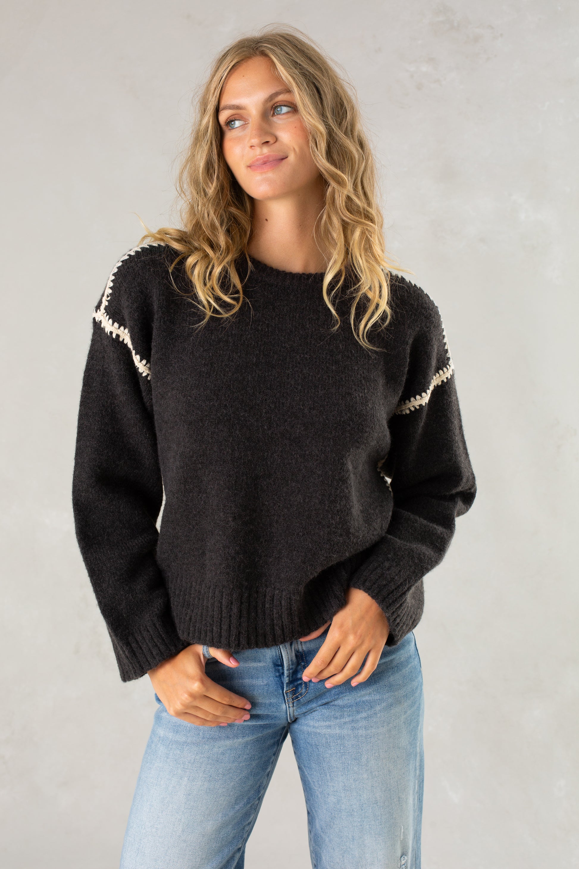A young woman with wavy blonde hair wears The Jenny Sweater by Isla in black, featuring a relaxed fit and subtle crochet details on the shoulders, along with light blue jeans, standing against a plain light gray background and softly smiling.