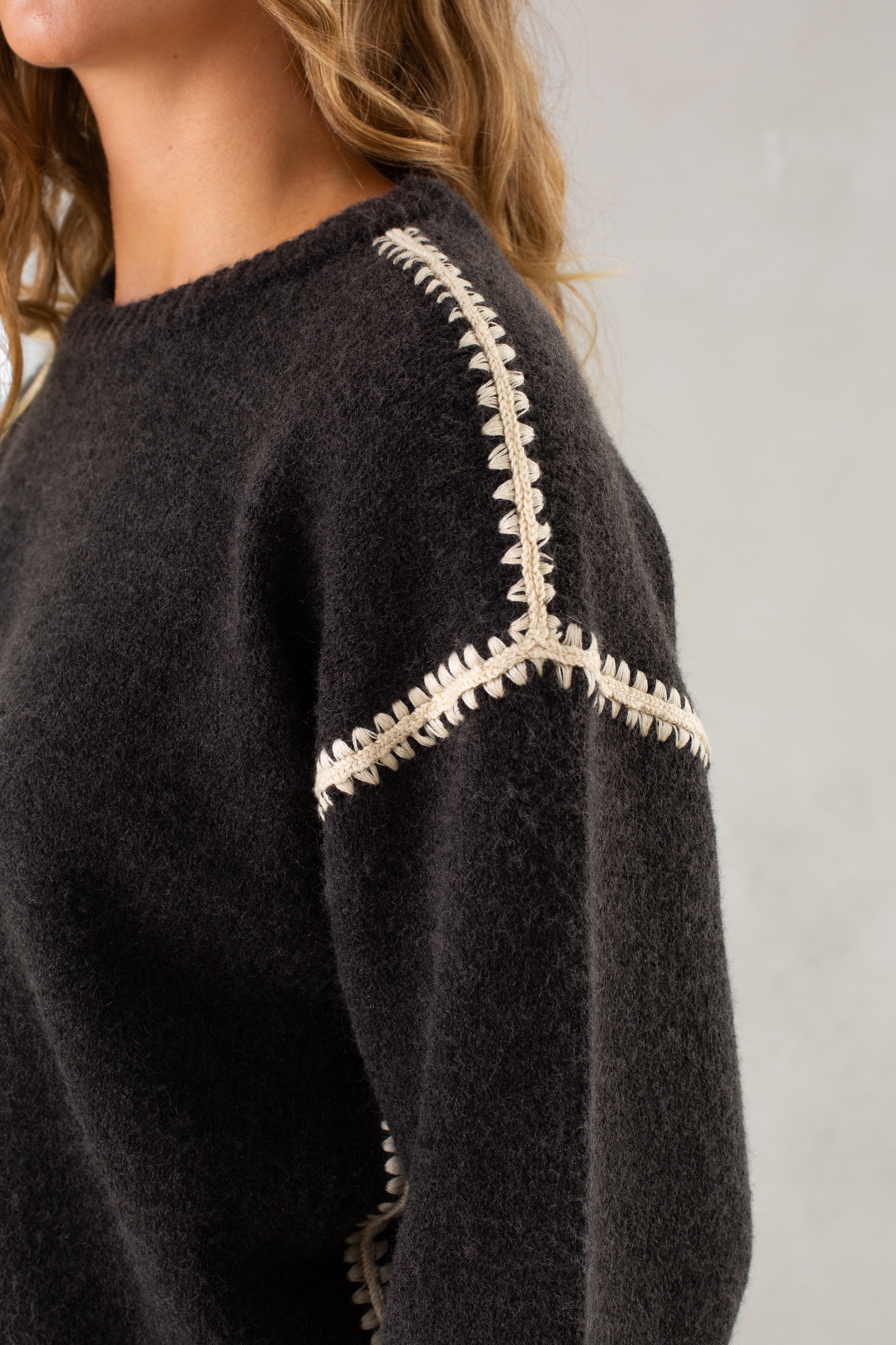 A person with wavy, light brown hair wears Isla’s The Jenny Sweater, a cozy crew neck with cream-colored stitched detailing along the shoulder and arm, set against a neutral background.