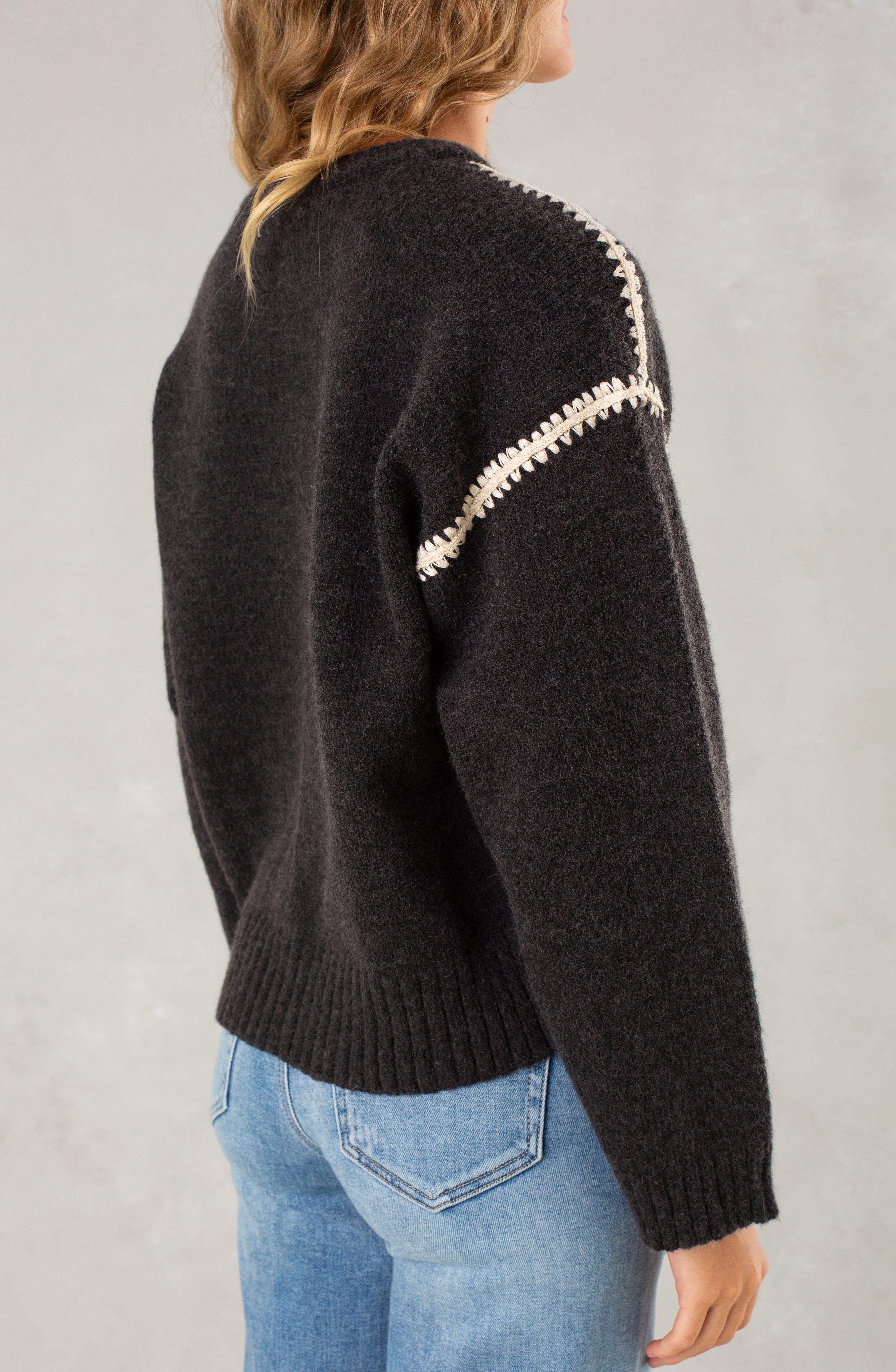 A person with wavy, shoulder-length hair faces away from the camera against a light background, wearing Isla's The Jenny Sweater—a relaxed-fit crew neck with crochet shoulder details—paired with light blue jeans.