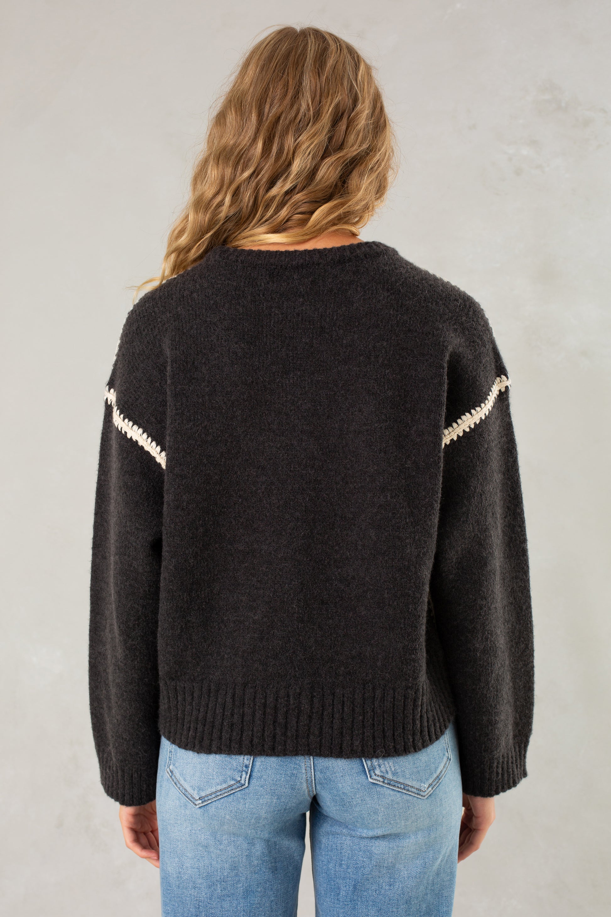 A person with wavy, light brown hair is shown from the back wearing Isla’s The Jenny Sweater, a relaxed-fit crew neck with white sleeve stripes, paired with light blue jeans against a light gray background.