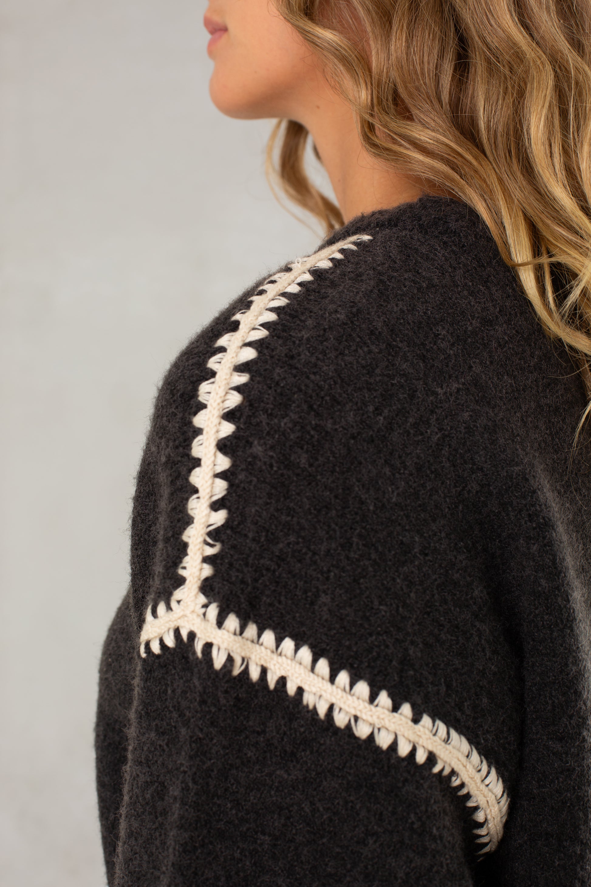A woman with wavy light brown hair wears Isla’s The Jenny Sweater, a relaxed-fit crew neck with cream crochet detailing on the shoulder and sleeve. Cropped side view emphasizes the sweater's unique texture.