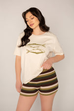 A woman with long dark hair wears the oversized boxy fit Fish Tee by Amanda Batula Swim, featuring two illustrated sardines, paired with green, brown, and cream striped shorts against a plain light background.