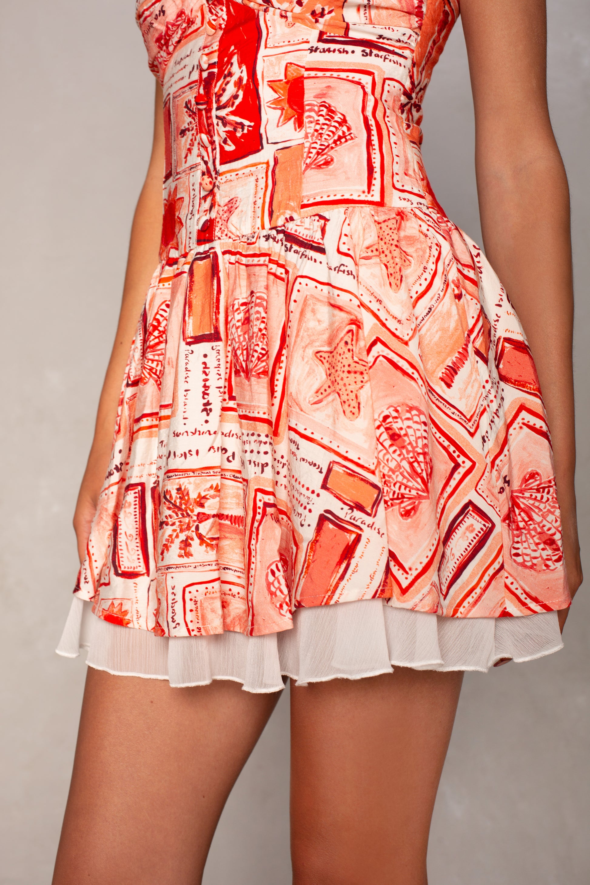 A person stands against a neutral background wearing the Isla Chloe Slim Mini Dress, a sleeveless mini featuring a coastal-inspired red and white abstract pattern with geometric and organic shapes, plus a flowy skirt and white ruffled hem.