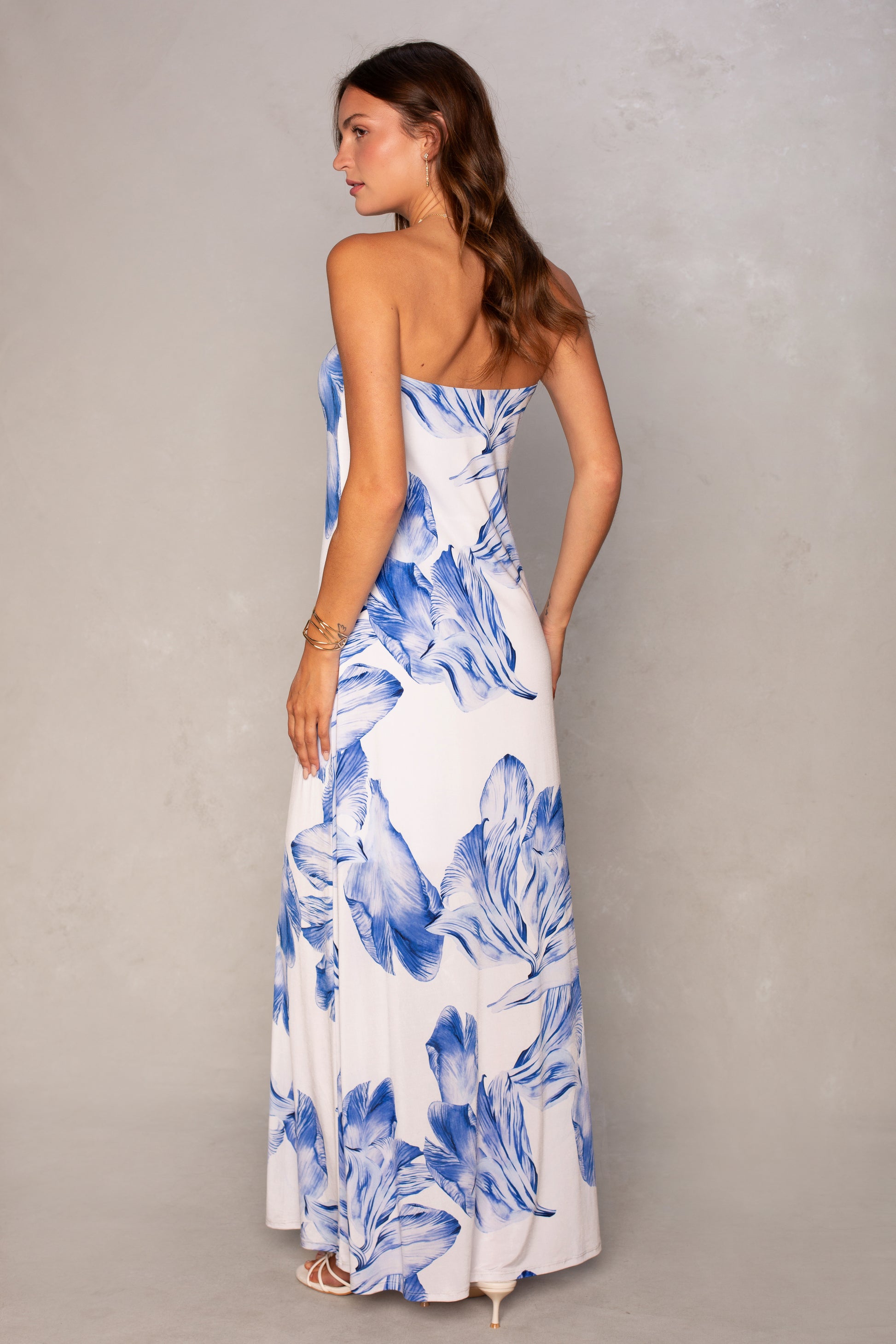 A woman in Isla's Flora Tube Maxi Dress, a strapless maxi with bold blue florals, stands looking over her shoulder. She wears white heels and a gold bracelet against a light gray backdrop—an ideal pick for summer events.