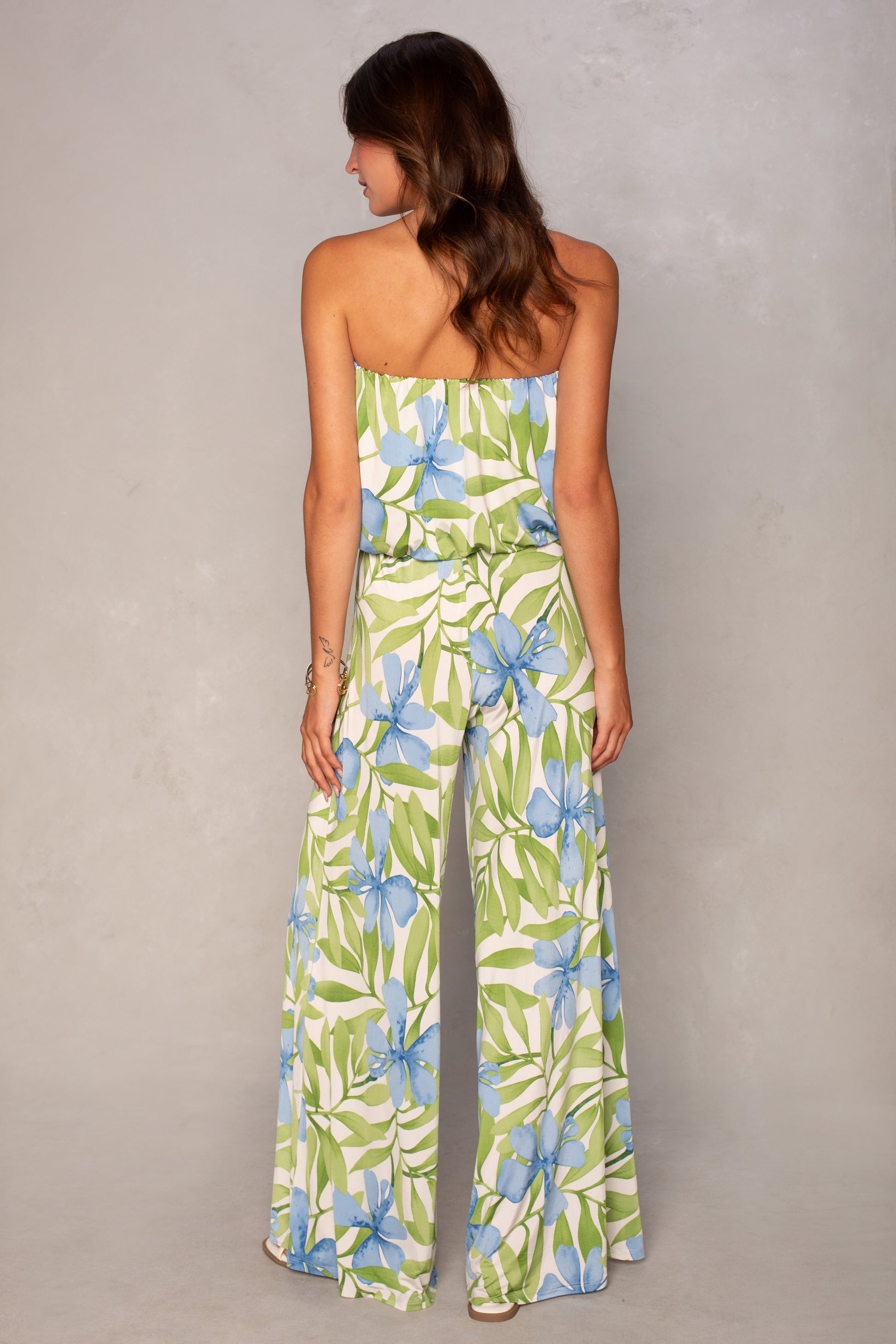 A woman with loose brown waves stands with her back to the camera in the Ari Strapless Easy Jumpsuit by Isla, featuring a tropical print of green leaves and blue flowers and wide leg pants.